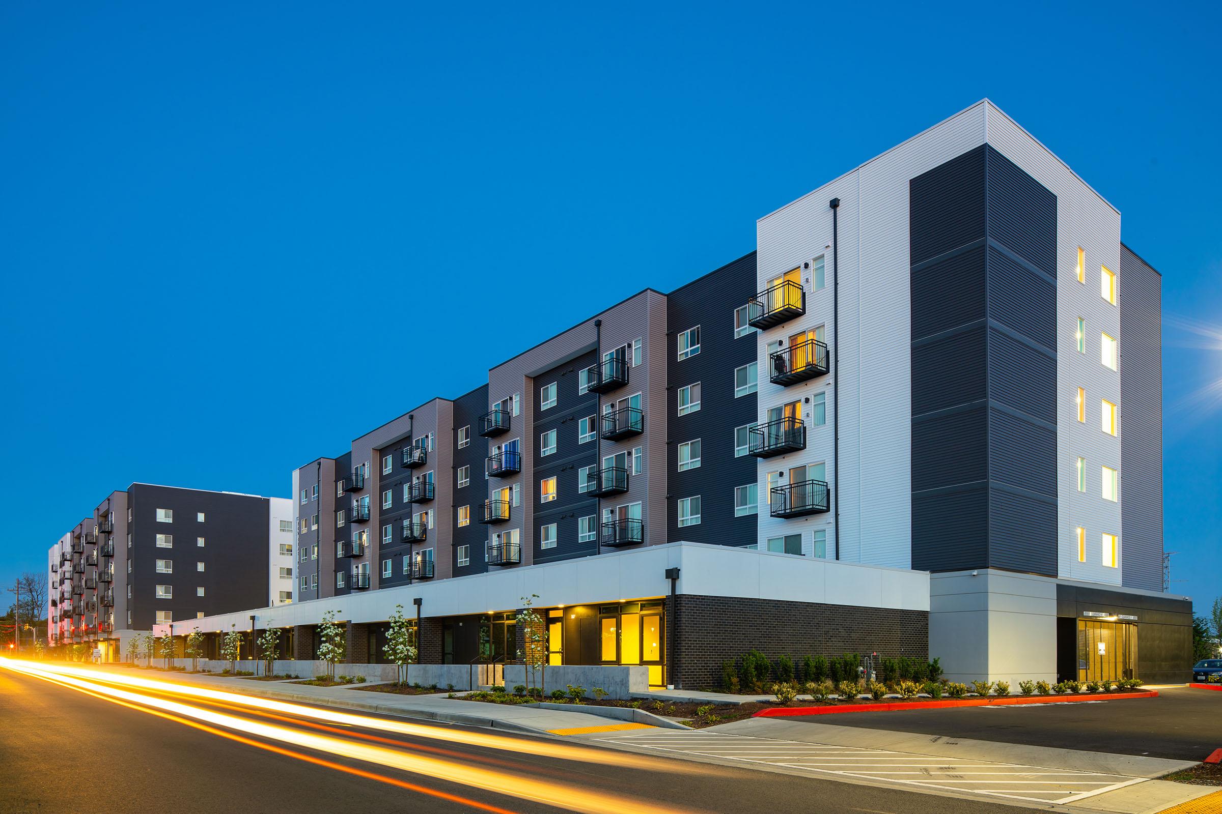 A modern multi-story apartment building with a mix of dark and light-colored exterior, featuring balconies and large windows. The scene is set during twilight, with car headlights creating streaks of light on the road in front of the building.