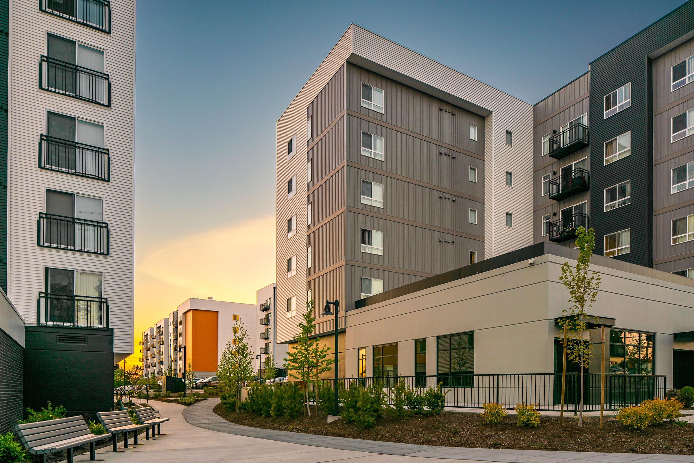 Modern apartment complex with three buildings, featuring balconies, large windows, and a landscaped pathway with benches. The scene is illuminated by a warm sunset sky.