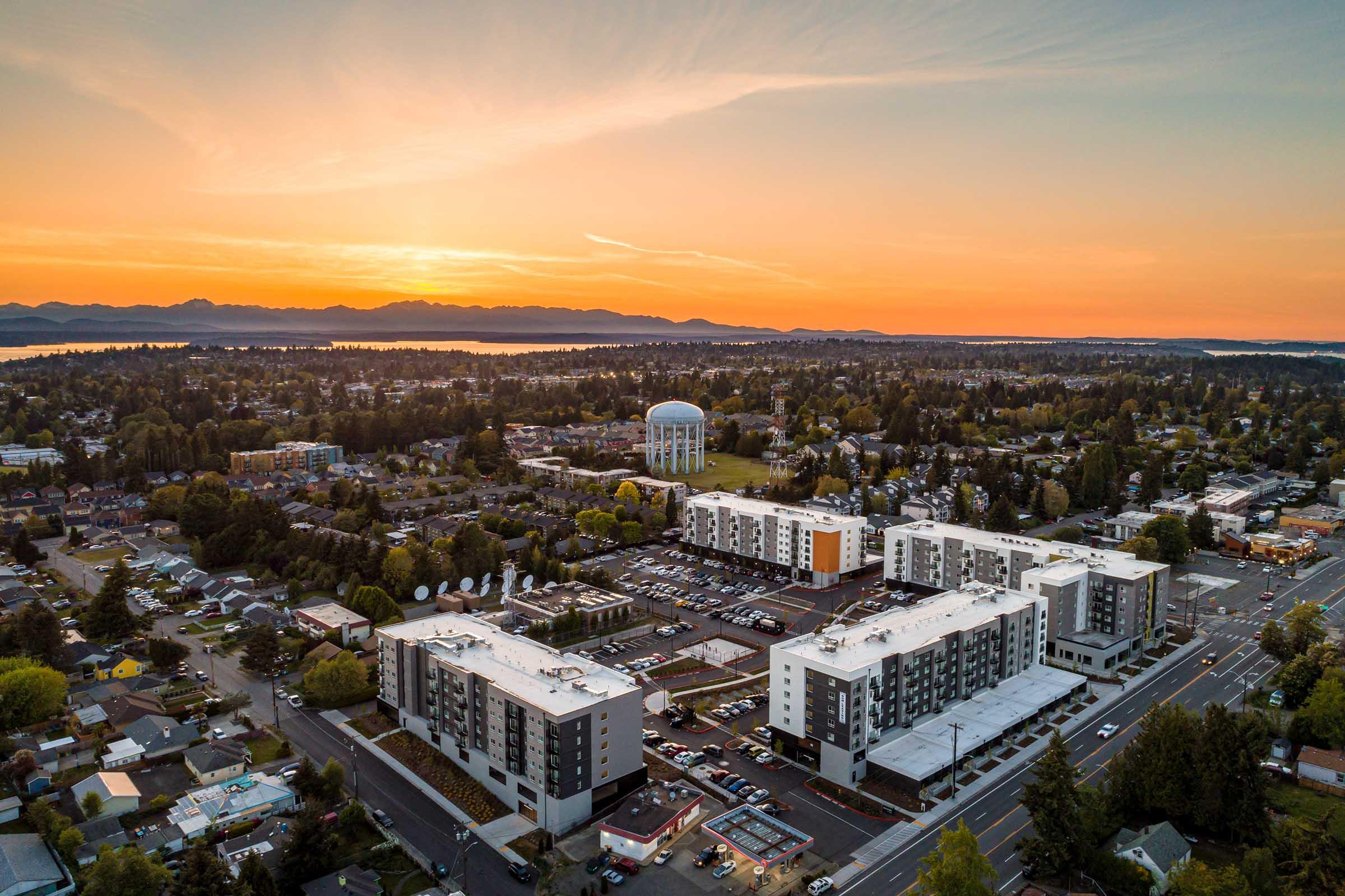 Aerial view of a suburban area at sunset, featuring a cluster of residential buildings, a water tower, and the horizon lined with mountains. Trees and a body of water are visible in the background, along with parking lots and roads.