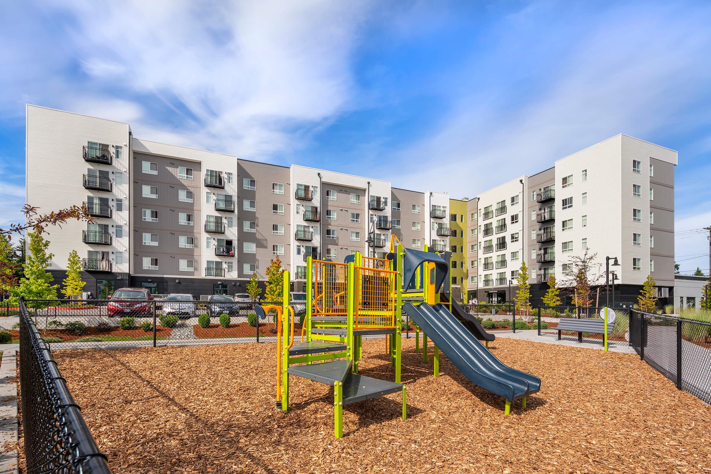 A playground featuring a yellow play structure with slides, surrounded by wood chips, in front of modern apartment buildings under a blue sky.