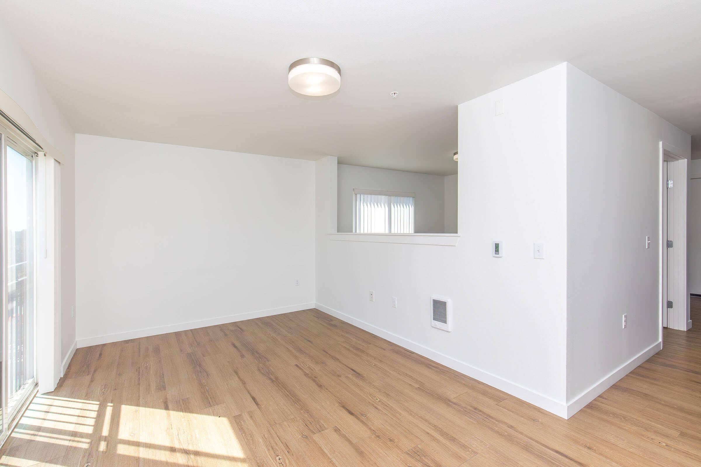 Bright and airy room with light-colored walls and wooden flooring. There are large windows allowing natural light, a ceiling light fixture, and an open layout leading to another space. The room appears empty and ready for furniture arrangement.