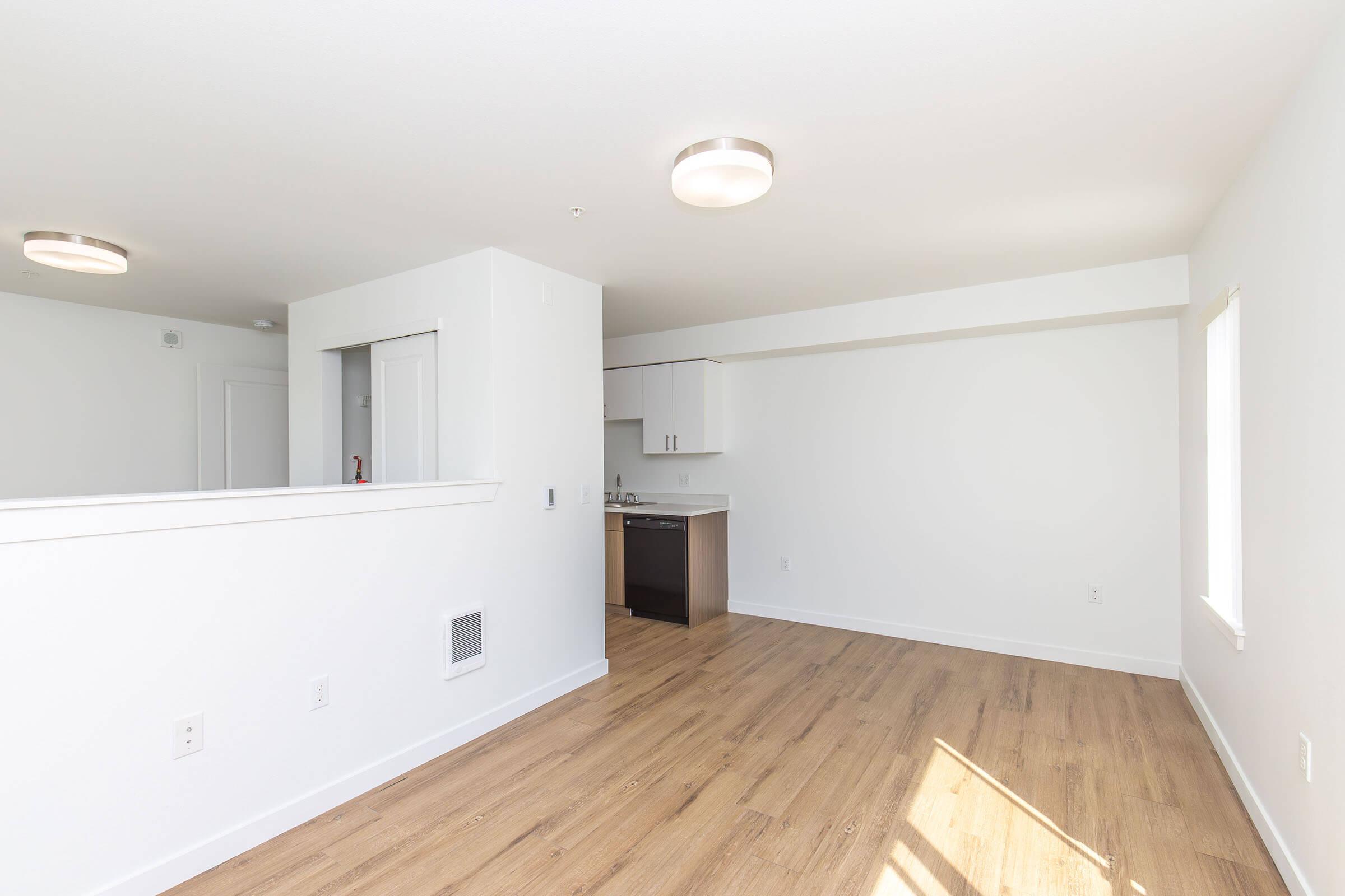 Bright and modern apartment interior featuring light wood flooring, white walls, and a spacious layout. The living area connects to a kitchen with white cabinetry, while natural light enters through a window. Two circular ceiling lights provide illumination, enhancing the contemporary feel of the space.