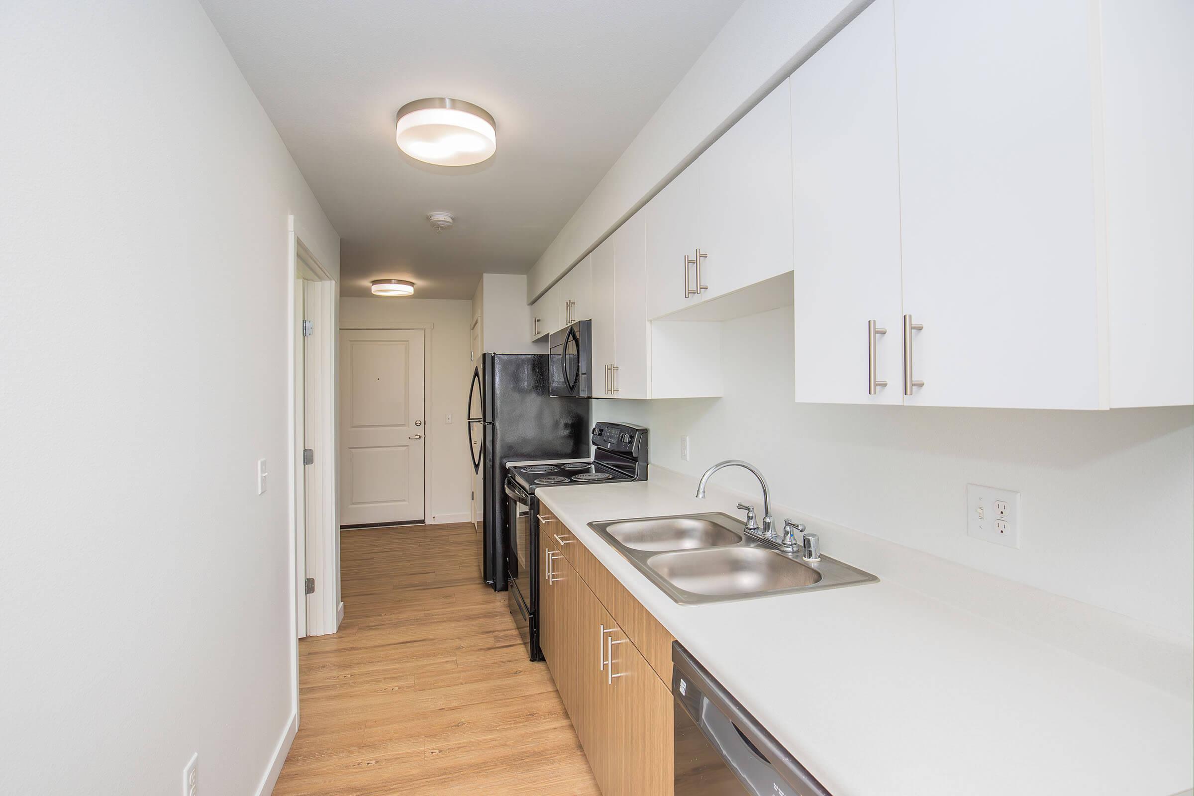 A modern kitchen with white cabinets, stainless steel appliances, and dual sinks. The space features a light wood floor and a door leading to a hallway. Bright overhead lighting illuminates the area, creating a clean and inviting atmosphere.