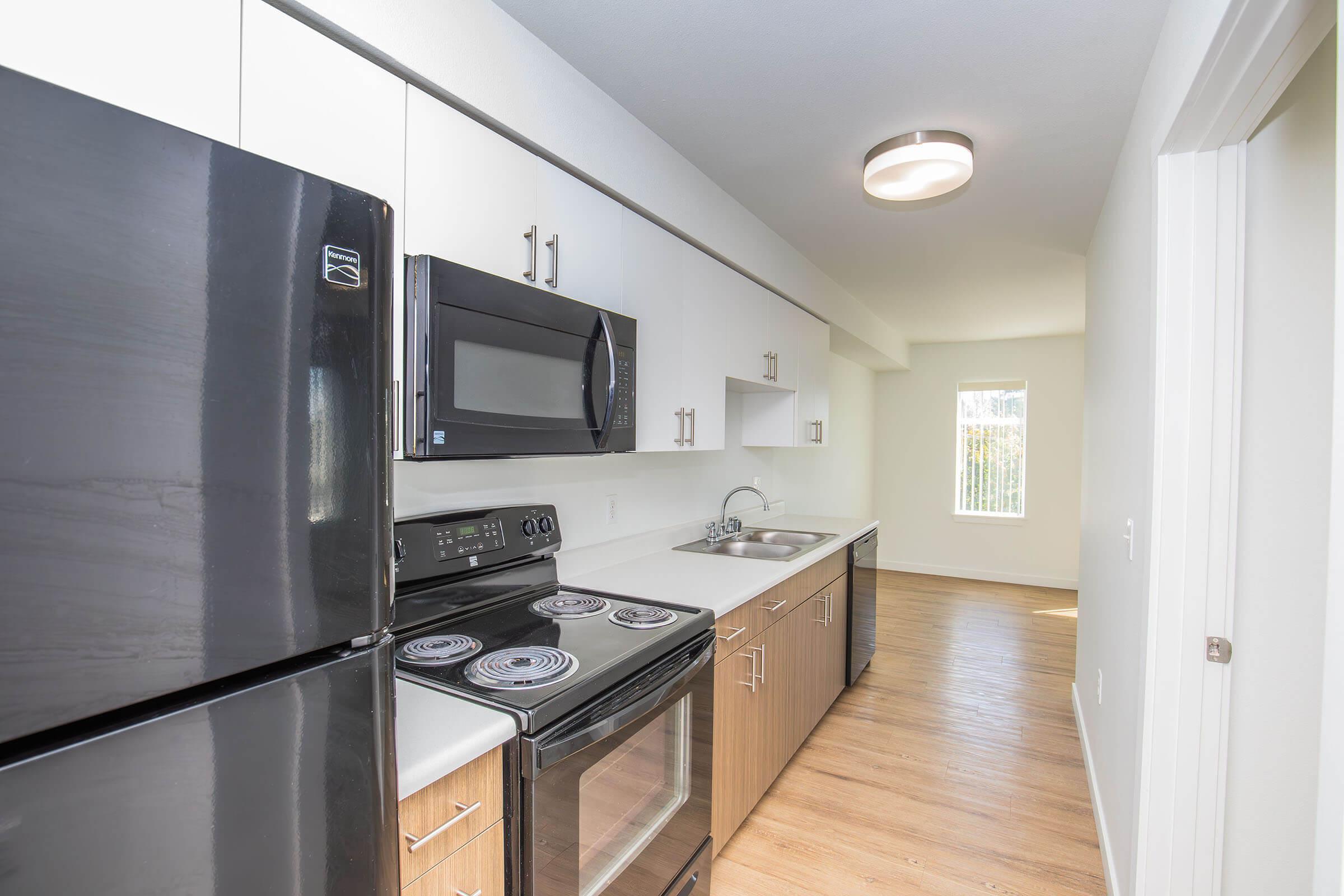 Modern kitchen featuring a black refrigerator, microwave, and electric stove. The countertops are white with a stainless steel sink. Light wood flooring extends into the background, where there's a window allowing natural light to enter the space. Sleek cabinetry complements the contemporary design.