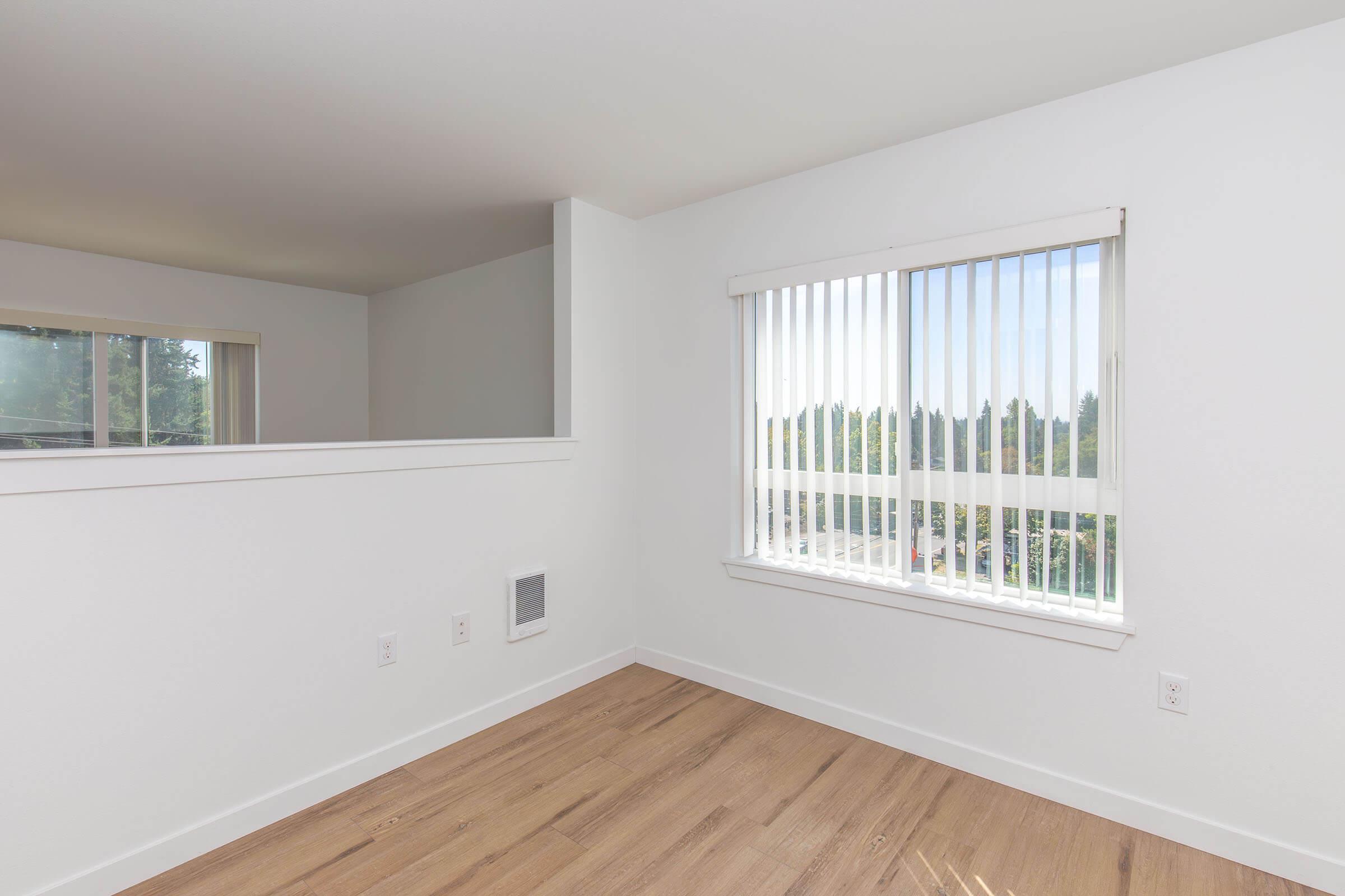 A bright and airy room with light-colored walls and wooden flooring. The window has white vertical blinds, allowing natural light to flow in. There is a small wall section separating the room from another area, and the view outside shows greenery.