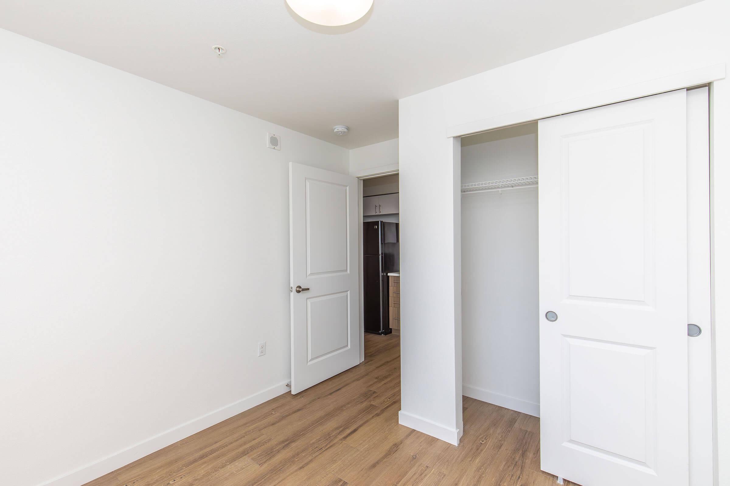 A bright, minimalist room featuring white walls and a light wood floor. To the right, there is a closet with sliding doors, and on the left, a white door leads to another space. Natural light illuminates the room, creating a clean and modern atmosphere.