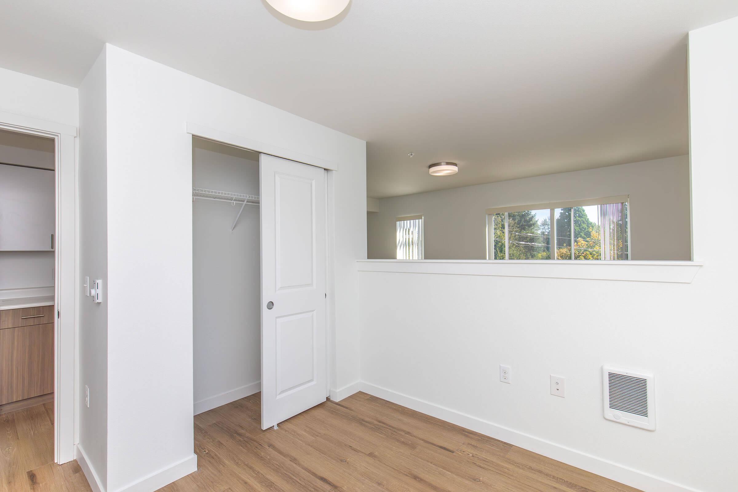 Interior view of a spacious room featuring light-colored walls, a wooden floor, and a closet with sliding doors. Natural light enters through a large window, and the design includes a dividing wall with a small opening. The room has a modern, minimalist aesthetic.