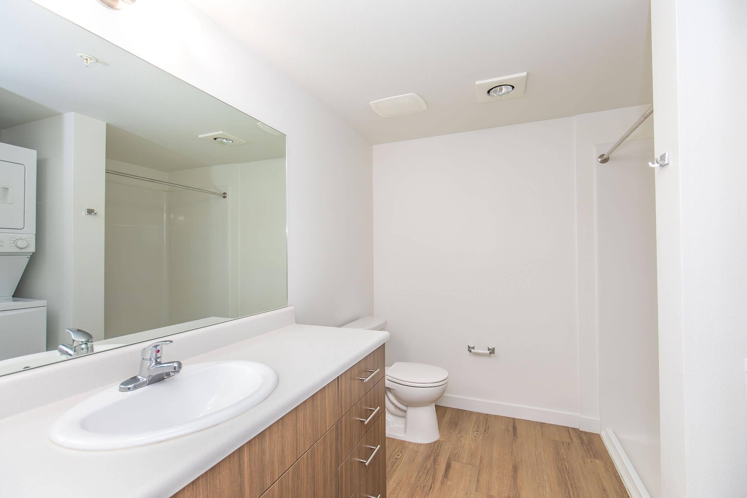 A clean and modern bathroom featuring a white countertop with a sink, a large mirror, wooden storage drawers, a toilet, and a shower area. The flooring is light wood, and there is a small space for a washing machine and dryer in the corner. The walls are painted white, creating a bright and spacious feel.