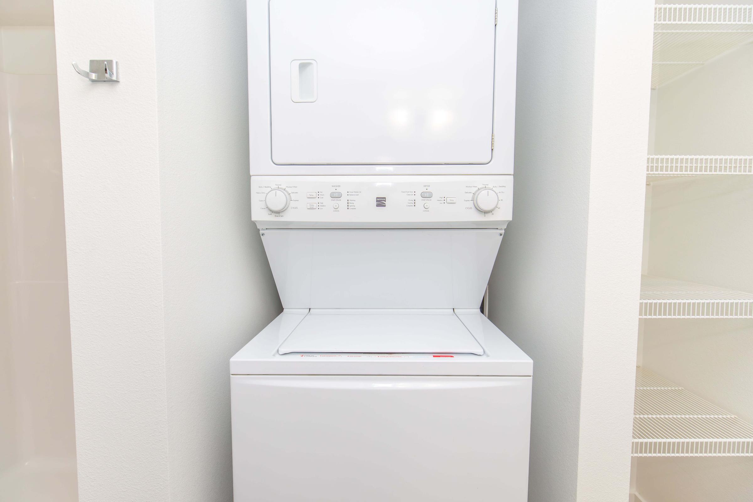 Stacked washer and dryer in a laundry nook. The white appliances are compact and feature control knobs and a door. The background has plain walls and shelving, providing a tidy and organized appearance.
