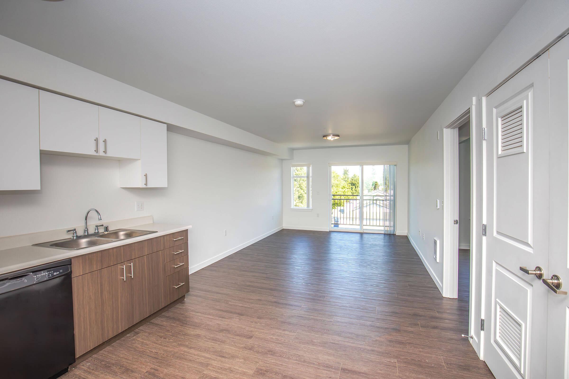 A modern, spacious kitchen and living area featuring light-colored walls, laminate flooring, a sink, and cabinetry. A large sliding glass door leads to a balcony, allowing natural light to fill the space. The room is unfurnished, creating a clean slate for decor.