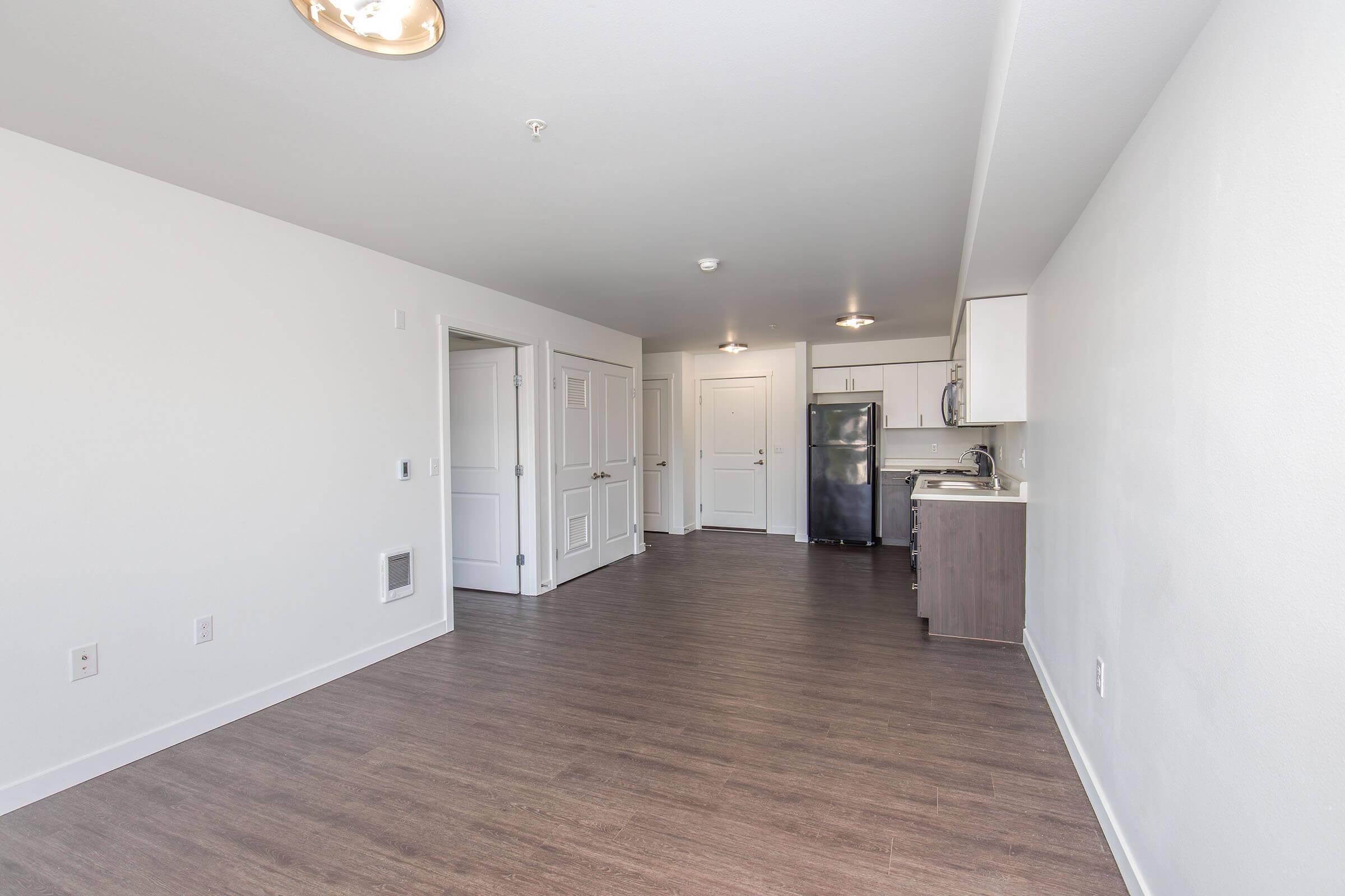 A spacious, modern apartment interior featuring a light-colored wall, hardwood flooring, and an open layout. The kitchen is equipped with stainless steel appliances, including a refrigerator. There are two doors leading to other rooms, and soft overhead lighting illuminates the space.