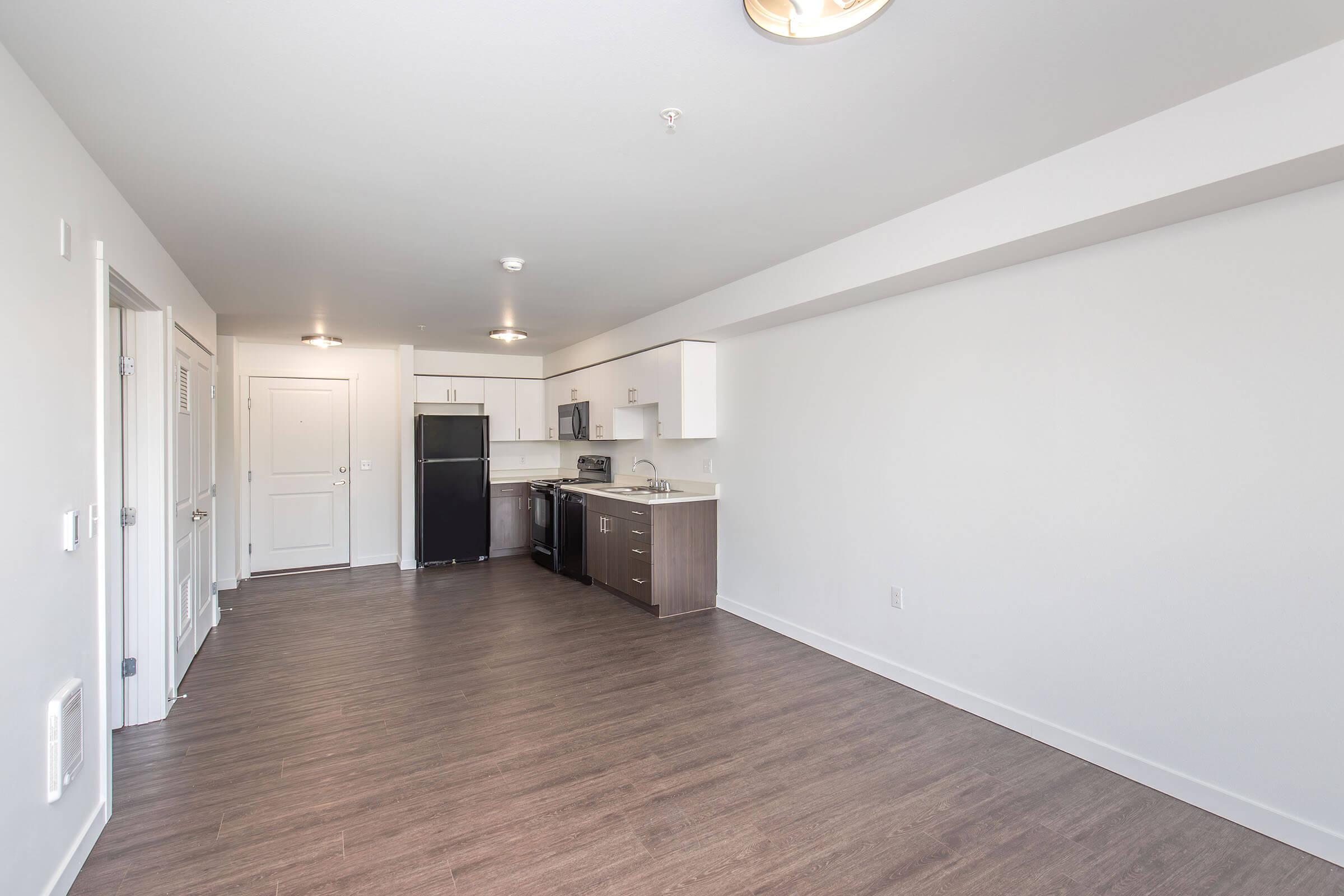 An empty modern apartment interior featuring an open kitchen and living area. The kitchen includes white cabinets, a black refrigerator, a stove, and a sink. The walls are painted light gray, and the floor is a light brown wood laminate. Bright lighting fixtures are installed on the ceiling.