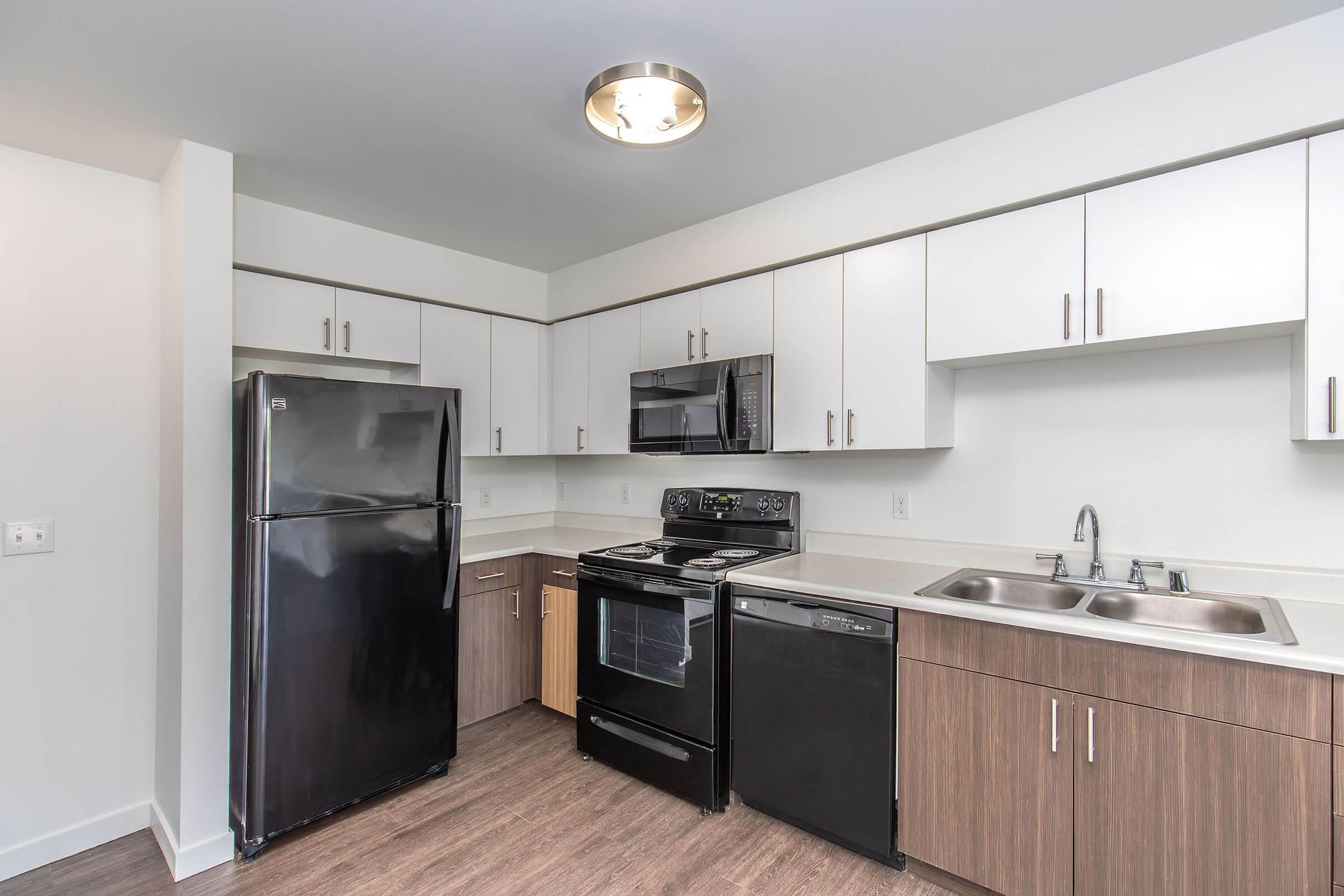 Modern kitchen featuring sleek black appliances, including a refrigerator, oven, and microwave. The cabinetry is a mix of white and wood finishes, providing a contemporary look. A double sink is installed in the countertop, with ample space and a light fixture above. The flooring is a light wood texture.