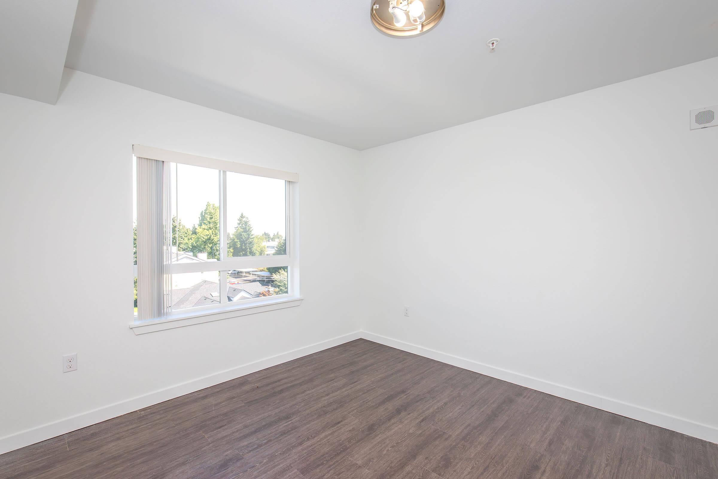 A bright, empty room featuring white walls, a large window with a view of greenery outside, and a modern ceiling light fixture. The flooring is a light wood laminate, creating a clean and spacious atmosphere.