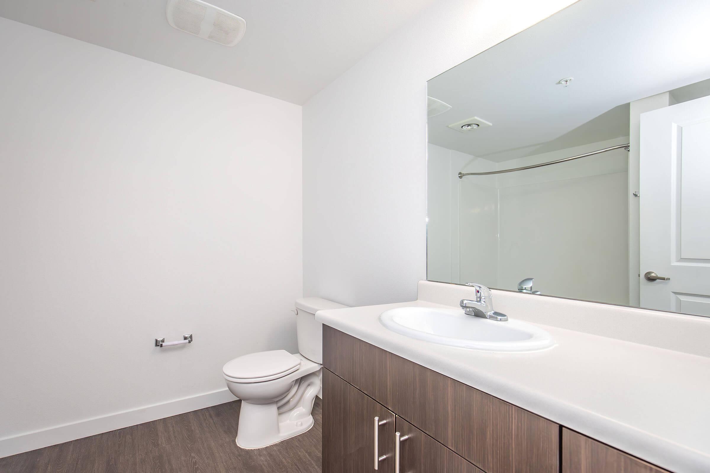 A modern bathroom featuring a toilet, a sink with a faucet, and a large mirror above the sink. The walls are painted white, and there is a shower area in the background with a curtain. The flooring is a wood-like material, creating a clean and minimalist aesthetic.