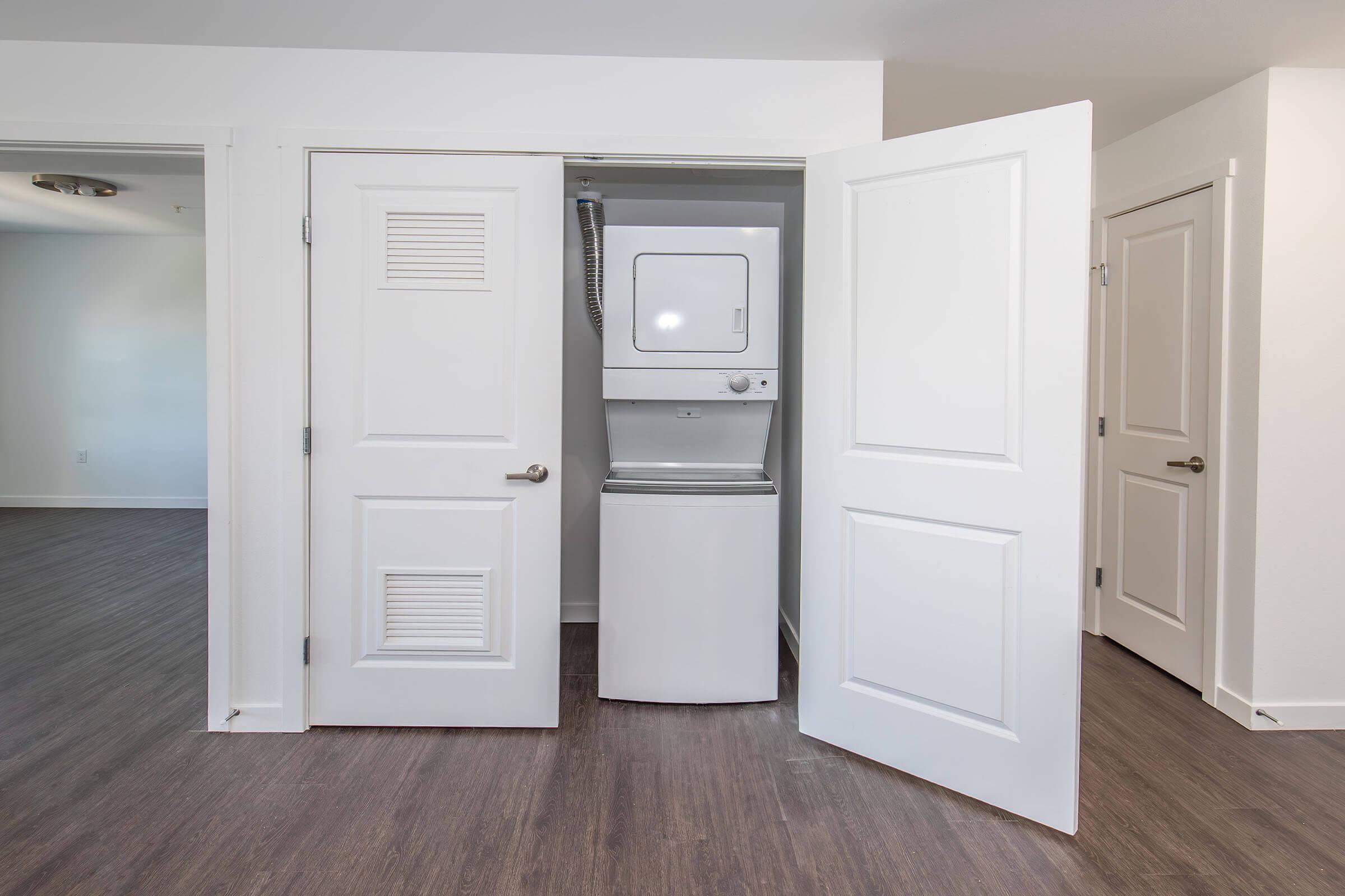 A laundry area featuring a stacked washer and dryer unit inside a closet. The closet has two white doors, which are open, revealing the appliances. The background shows a light-colored room with hardwood flooring and minimal furnishings.