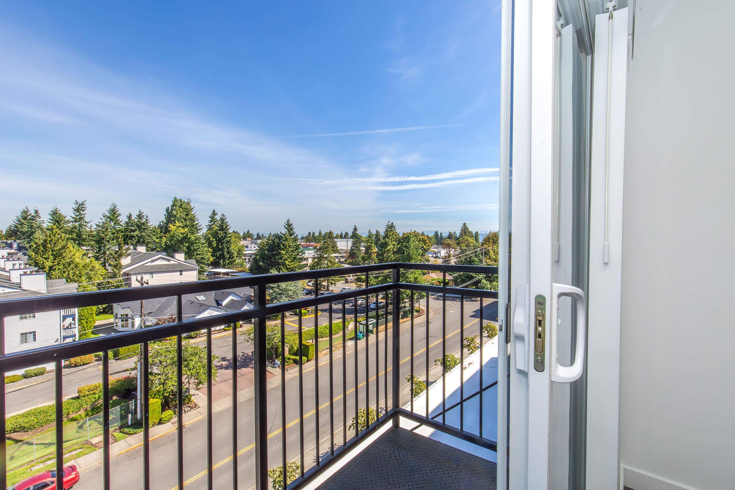 A balcony view overlooking a suburban neighborhood with trees, houses, and a clear blue sky. The balcony door is open, inviting natural light into the space.