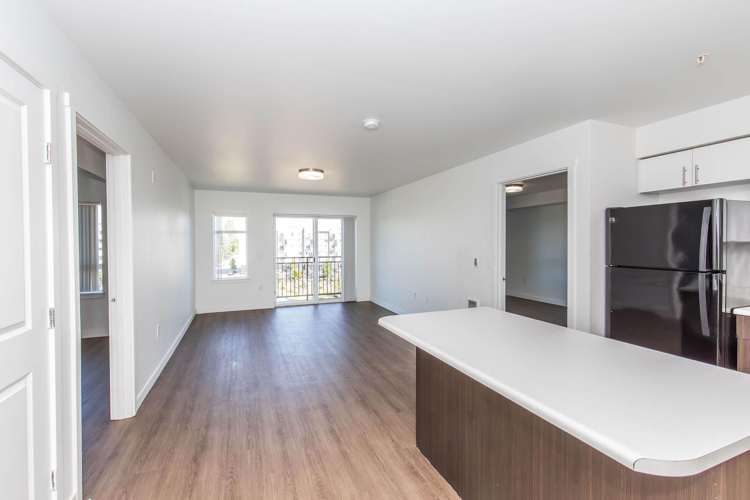 Spacious open-concept living area featuring a modern kitchen with an island, sleek black refrigerator, and large windows providing natural light. The room has a neutral color palette and hardwood flooring, leading to a balcony for outdoor access. Adjacent space visible for additional living or dining options.