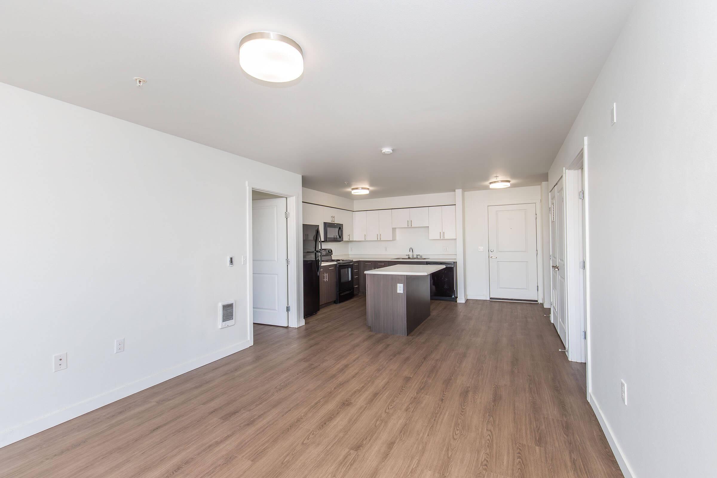 A modern, empty apartment interior featuring light-colored walls and wood-style flooring. The open kitchen includes an island and standard appliances. Doors lead to different areas, and bright overhead lighting enhances the spacious feel.