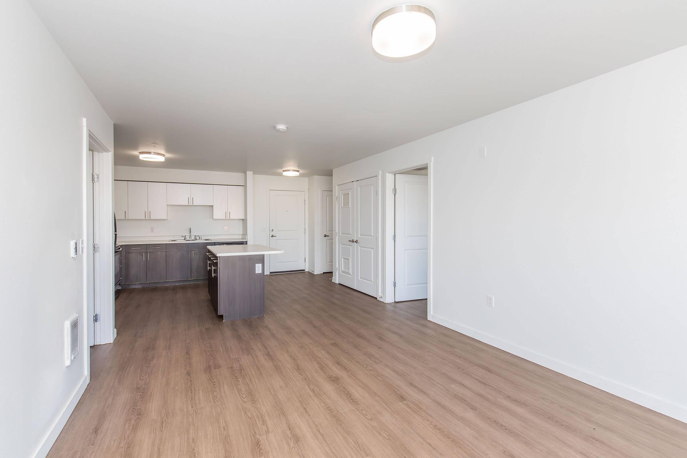 A modern interior of an empty apartment showing a living area with hardwood floors, a kitchen island, and kitchen cabinetry. The walls are painted white and there are doors leading to other rooms. Soft lighting fixtures are installed in the ceiling. The space is bright and spacious, ready for furnishing.