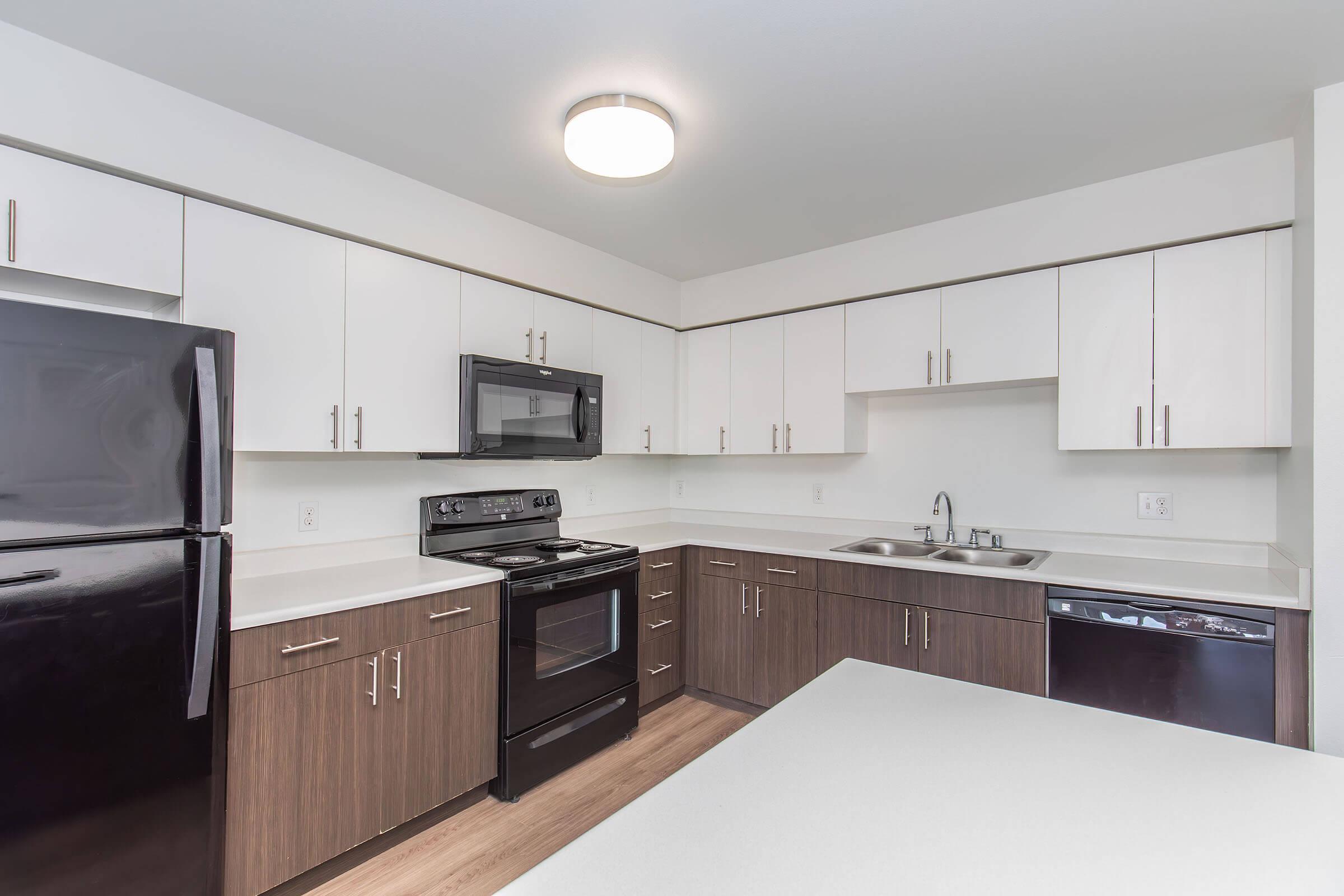 Modern kitchen with white and dark wood cabinets, a black refrigerator, stove, and microwave. The countertop is light-colored, and there are dual sinks beneath a window. The space features an open layout with a clean, minimalistic design.