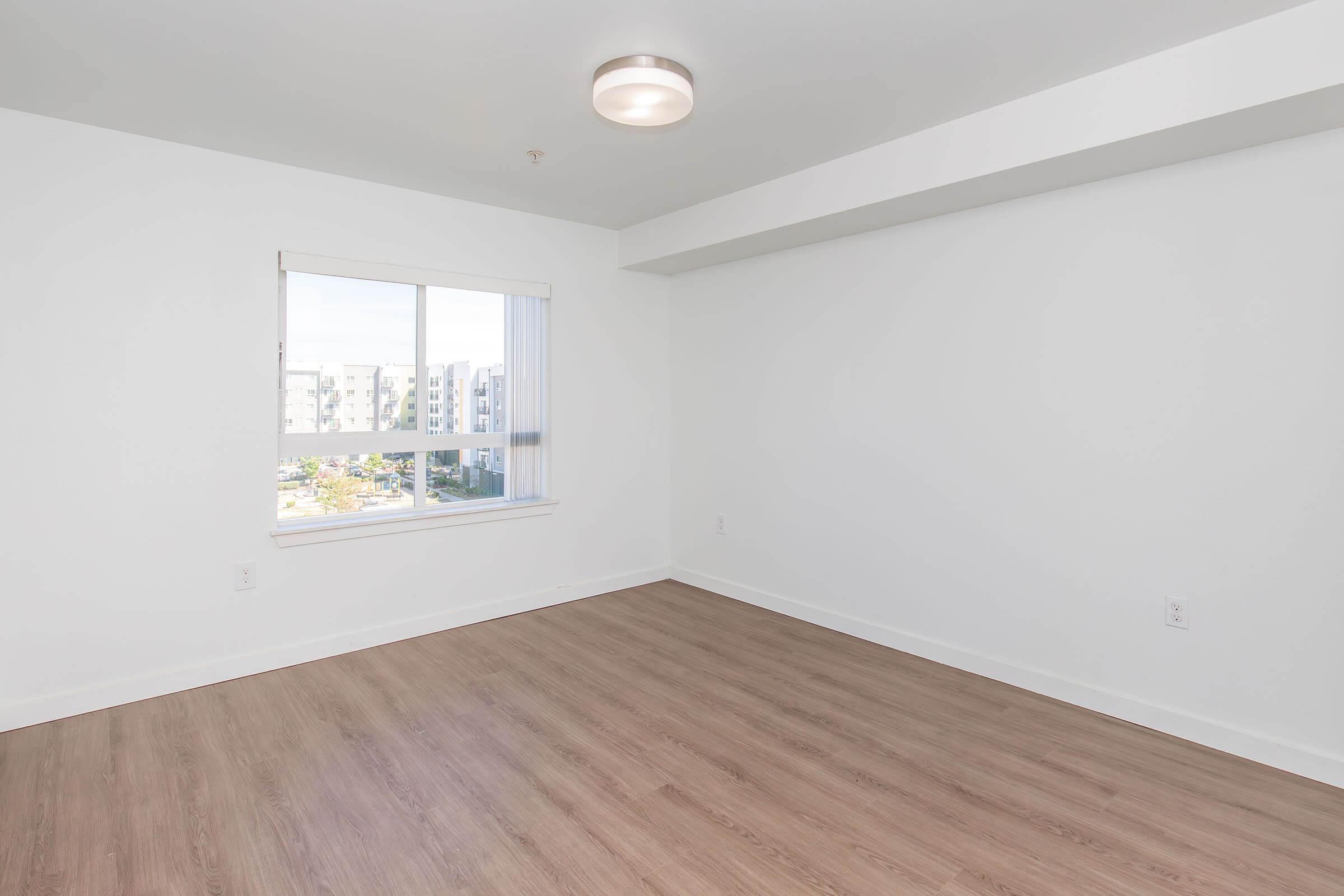 Empty room with light-colored walls and a window letting in natural light. The floor is a light wood laminate, and there is a modern ceiling light fixture. The view through the window shows an exterior with buildings in the background. The space is clean and minimalistic.