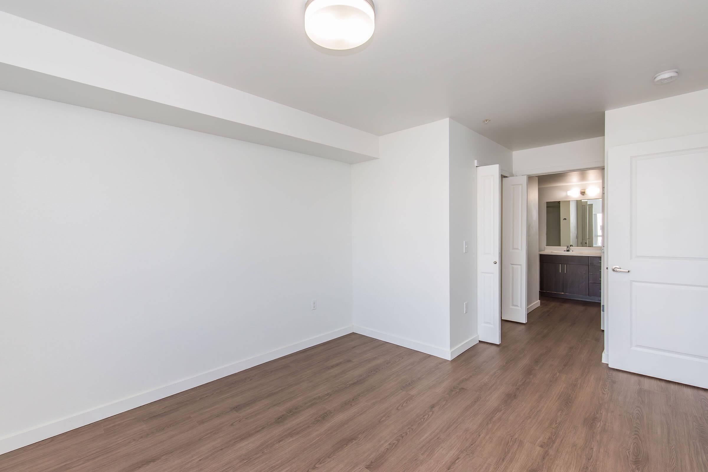Empty room with light-colored walls and wooden flooring. A doorway on the right leads to a small bathroom with a sink and mirror, visible from the main living space. A modern light fixture is installed on the ceiling. The room is spacious and bright, creating an inviting atmosphere.