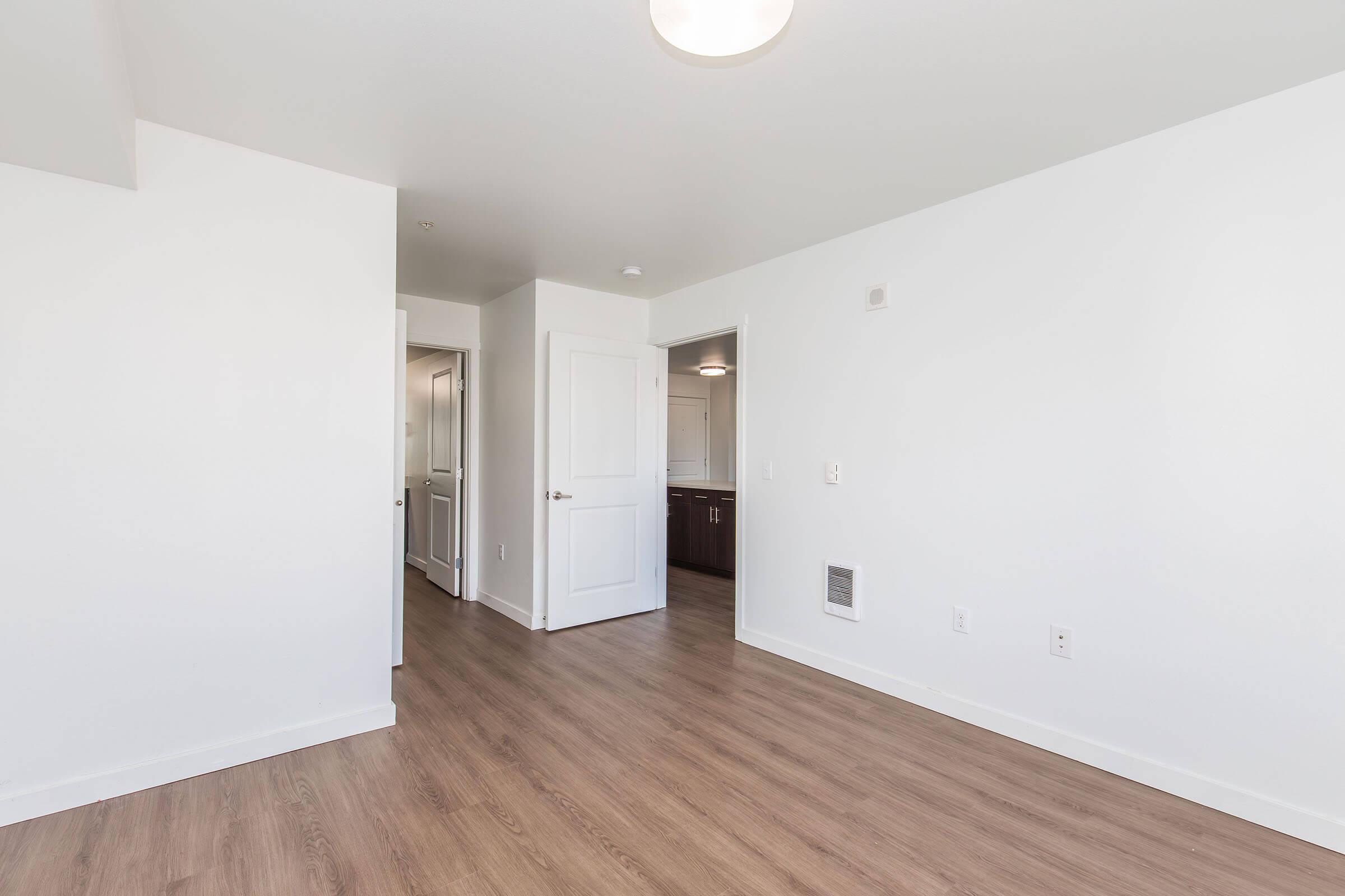 A bright, empty room featuring light-colored walls and wood-style flooring. The space includes a doorway leading to a kitchen area, a closet, and a door to another room. A ceiling light fixture is visible, enhancing the openness of the layout.