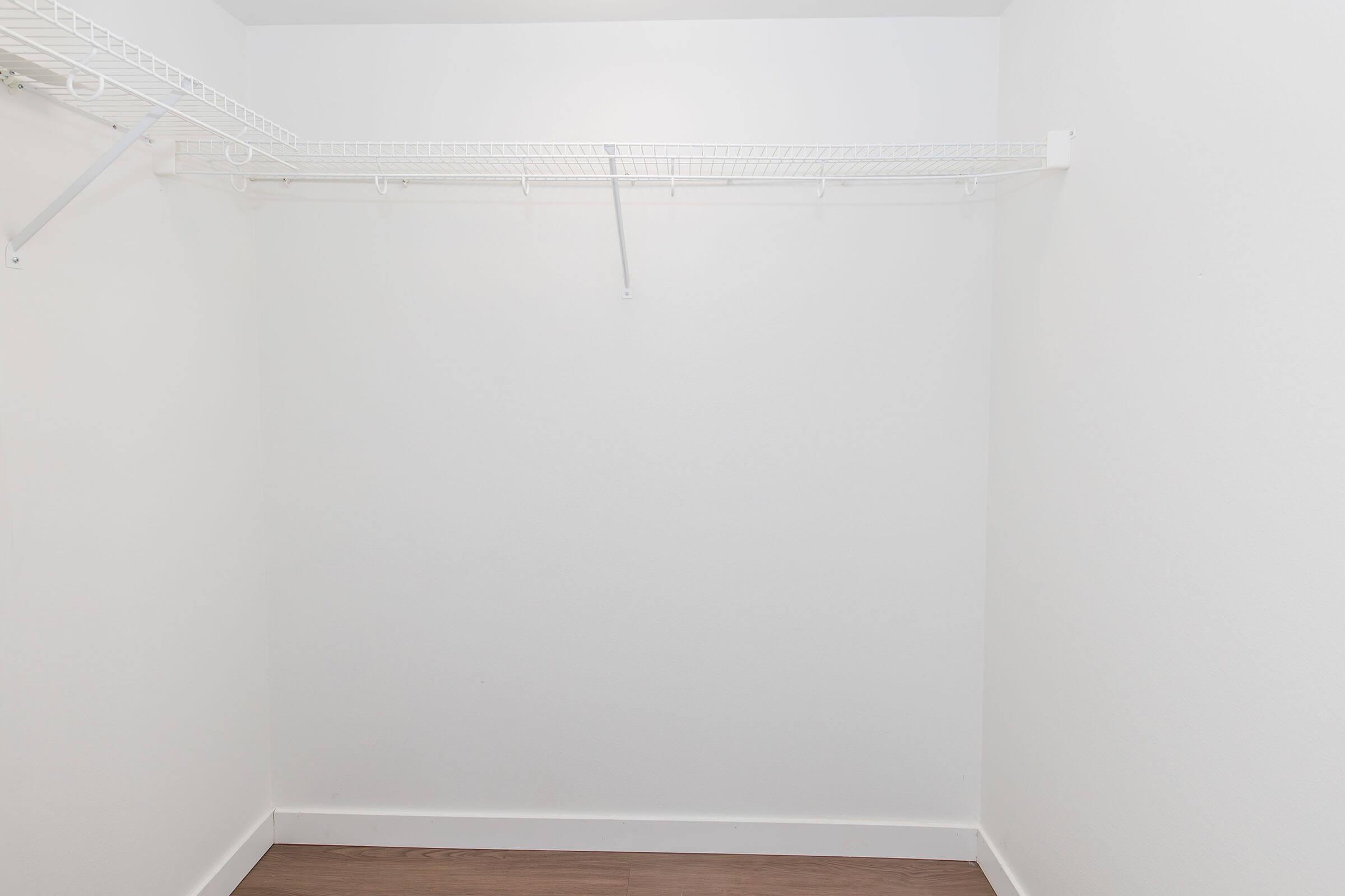 A minimally furnished closet with white walls and a wooden floor, featuring a wire shelving unit on the left side. The space is empty, with no items stored, creating an open and spacious appearance.