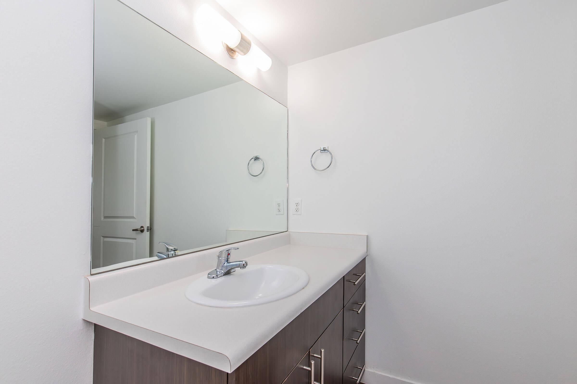 A clean, modern bathroom featuring a large mirror above a white sink with a faucet. The countertop is light-colored and smooth. There's a towel ring on the wall and dark wooden cabinetry beneath the sink. The walls are painted white, creating a bright and airy atmosphere.