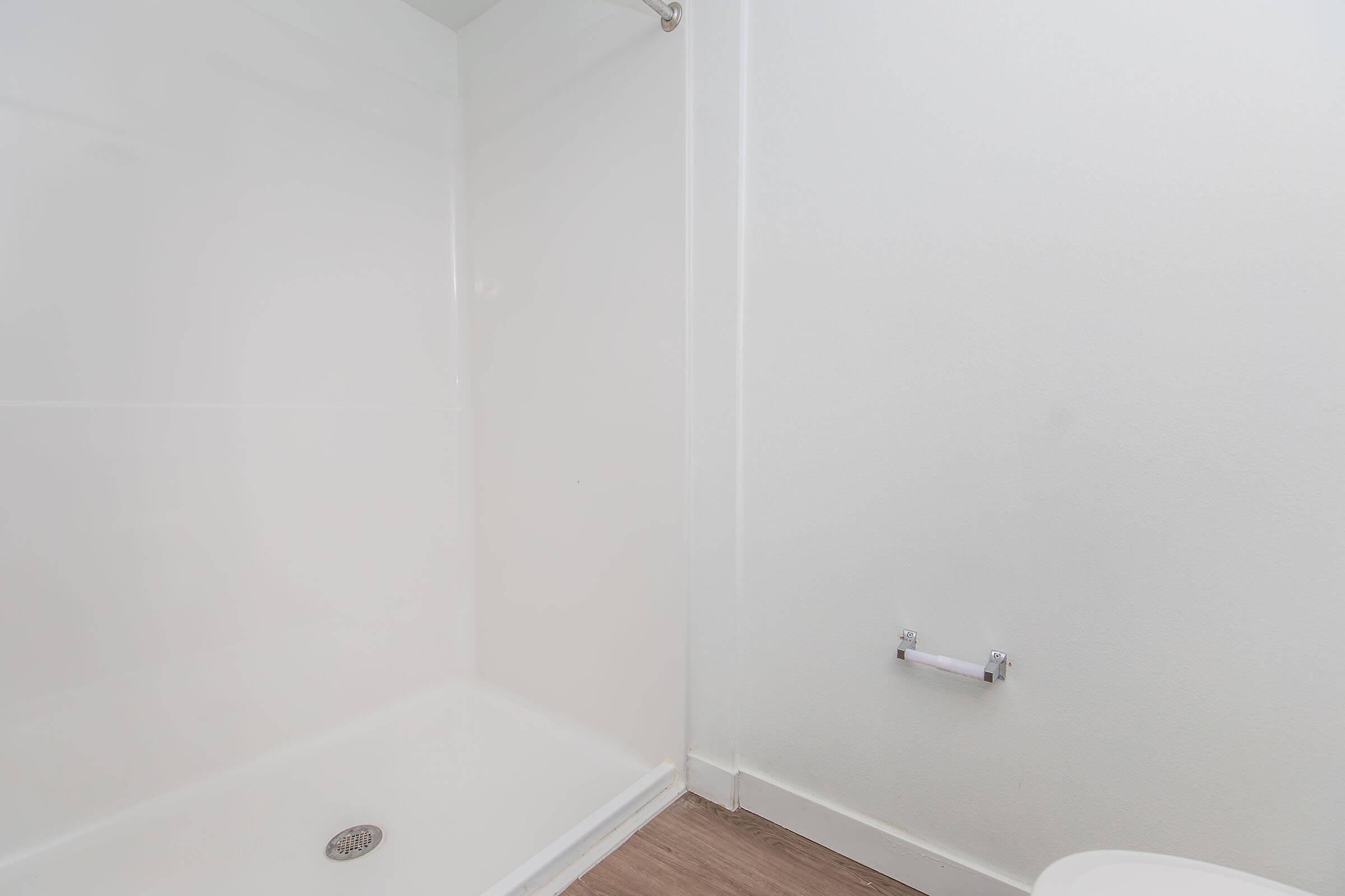A clean, white shower enclosure with a showerhead and a drain on the floor, adjacent to a wall with minimal decoration. A toilet paper holder is mounted on the wall nearby, and the flooring is wood-like. The overall atmosphere is bright and well-maintained.