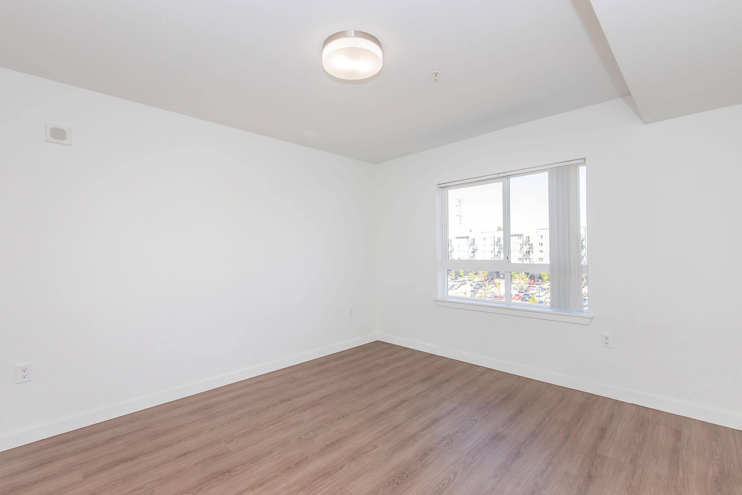 An empty room with white walls and a single window allowing natural light to enter. The floor is covered with light-colored wood laminate. A modern ceiling light fixture is installed in the center of the ceiling. The space appears spacious and clean, ready for furniture.