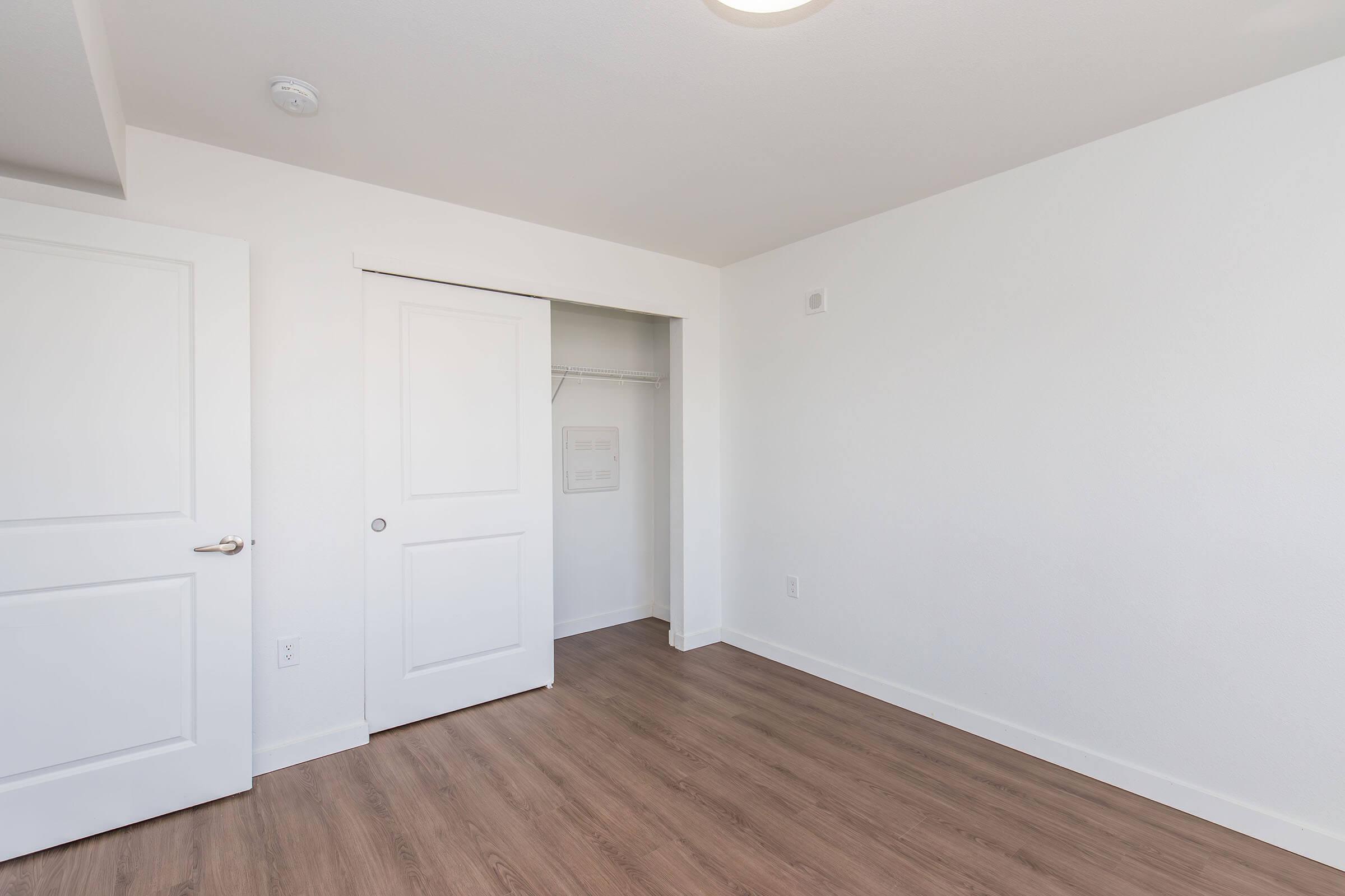 A clean, empty room featuring light hardwood flooring, white walls, and a closet with sliding doors. The room includes a ceiling light fixture and no furniture, creating a spacious, minimalist atmosphere.