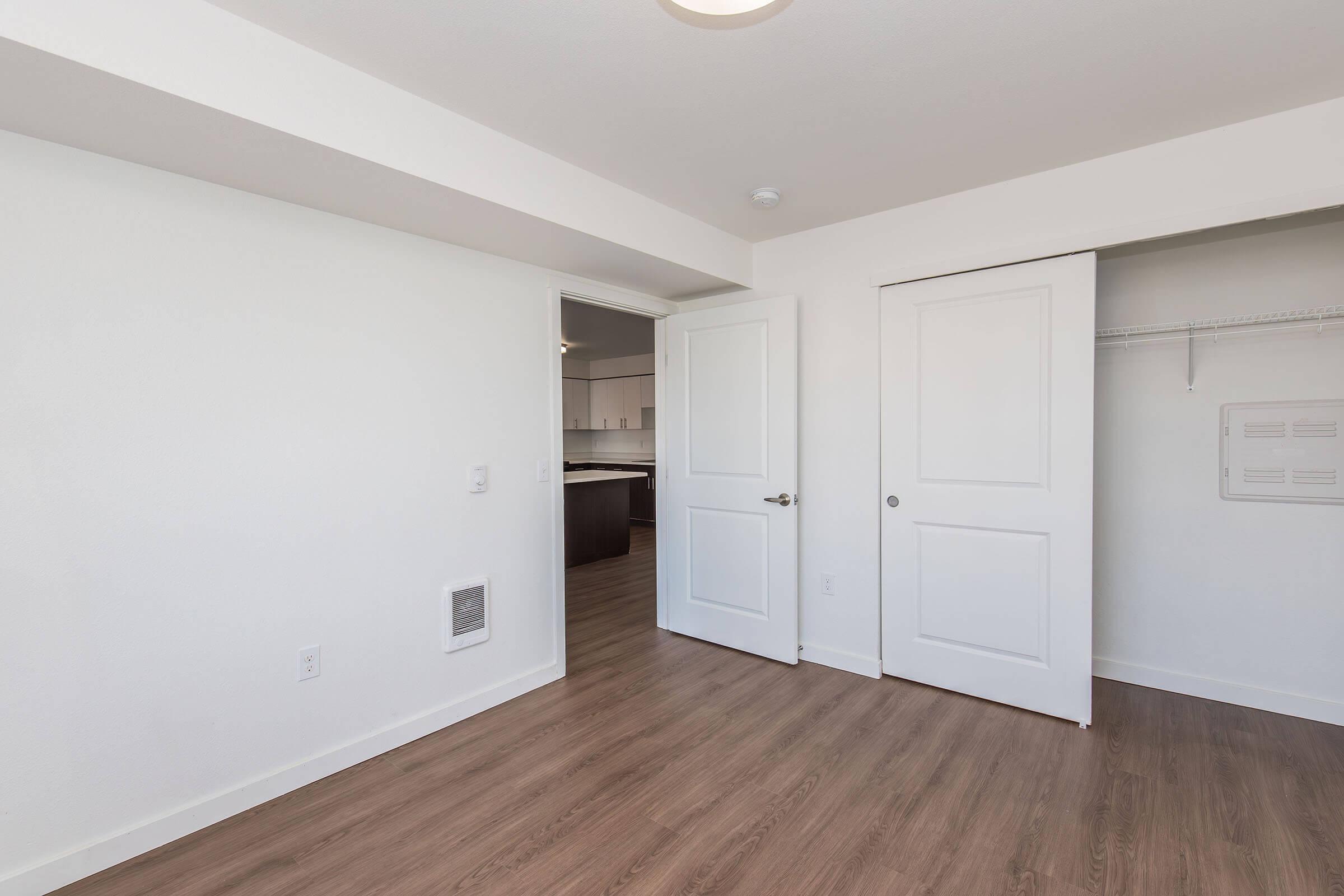 Empty interior room with light-colored walls and hardwood flooring. A door leads to an adjoining room, while another door opens to a closet. The room is well-lit with natural light and has a modern, minimalist design.