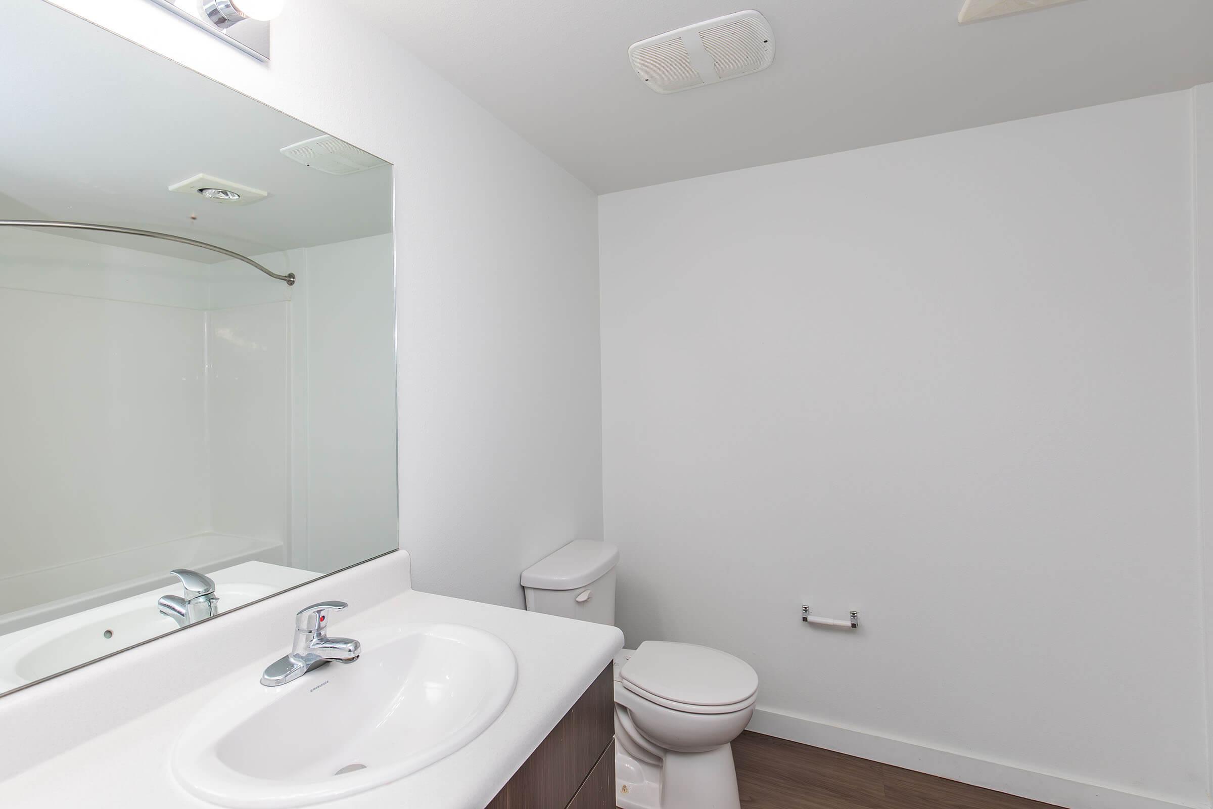 A clean, modern bathroom featuring a white sink with a faucet, a mirror above the sink, a white toilet, and a shower area with glass doors. The walls are painted white, and the flooring appears to be a light brown laminate. Natural light is coming from a window or overhead light source.