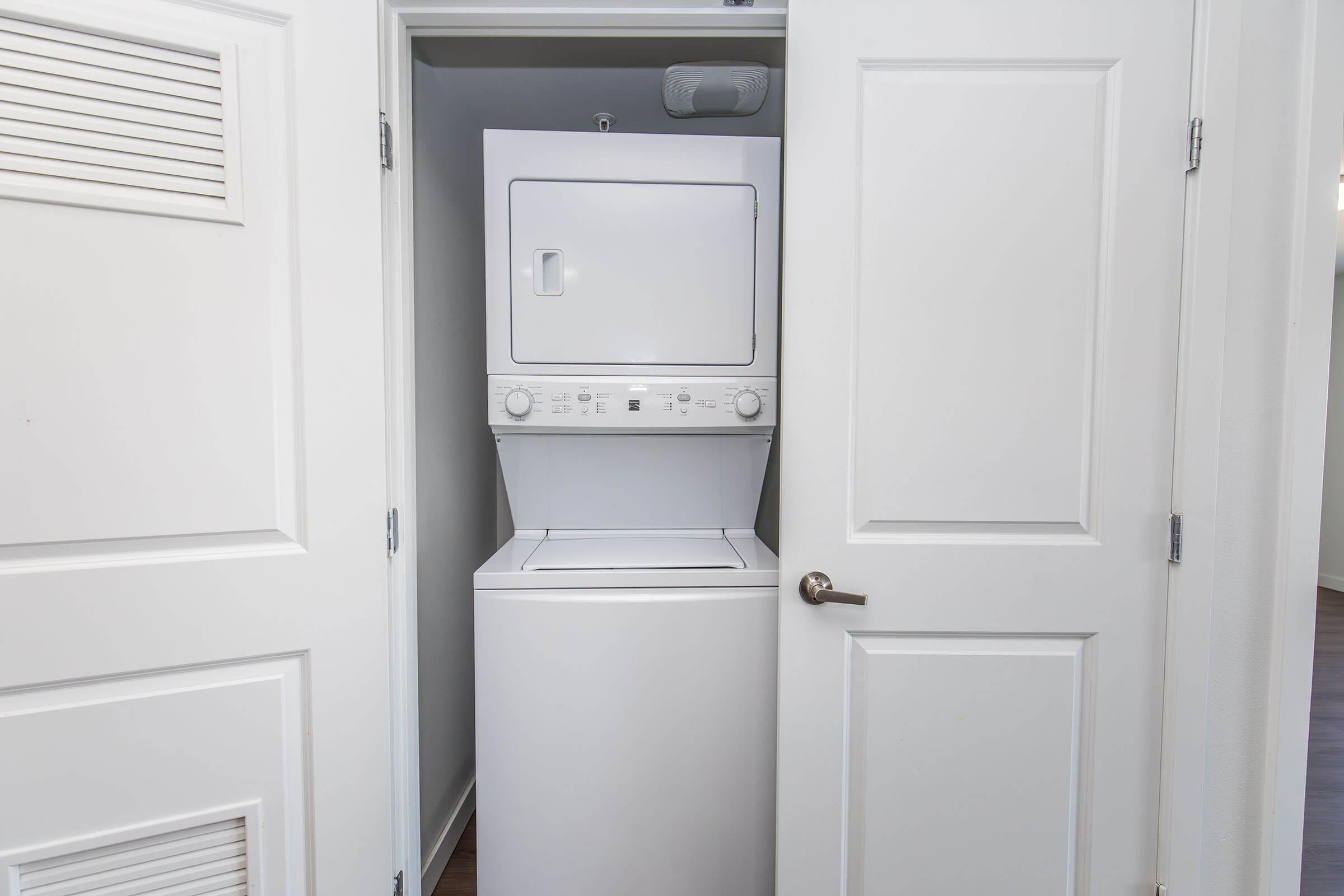 A stacked washer and dryer unit located in a closed closet with white doors. The appliances are white, featuring dials and buttons on the front. The interior of the closet is well-lit, showcasing clean lines and a modern design.