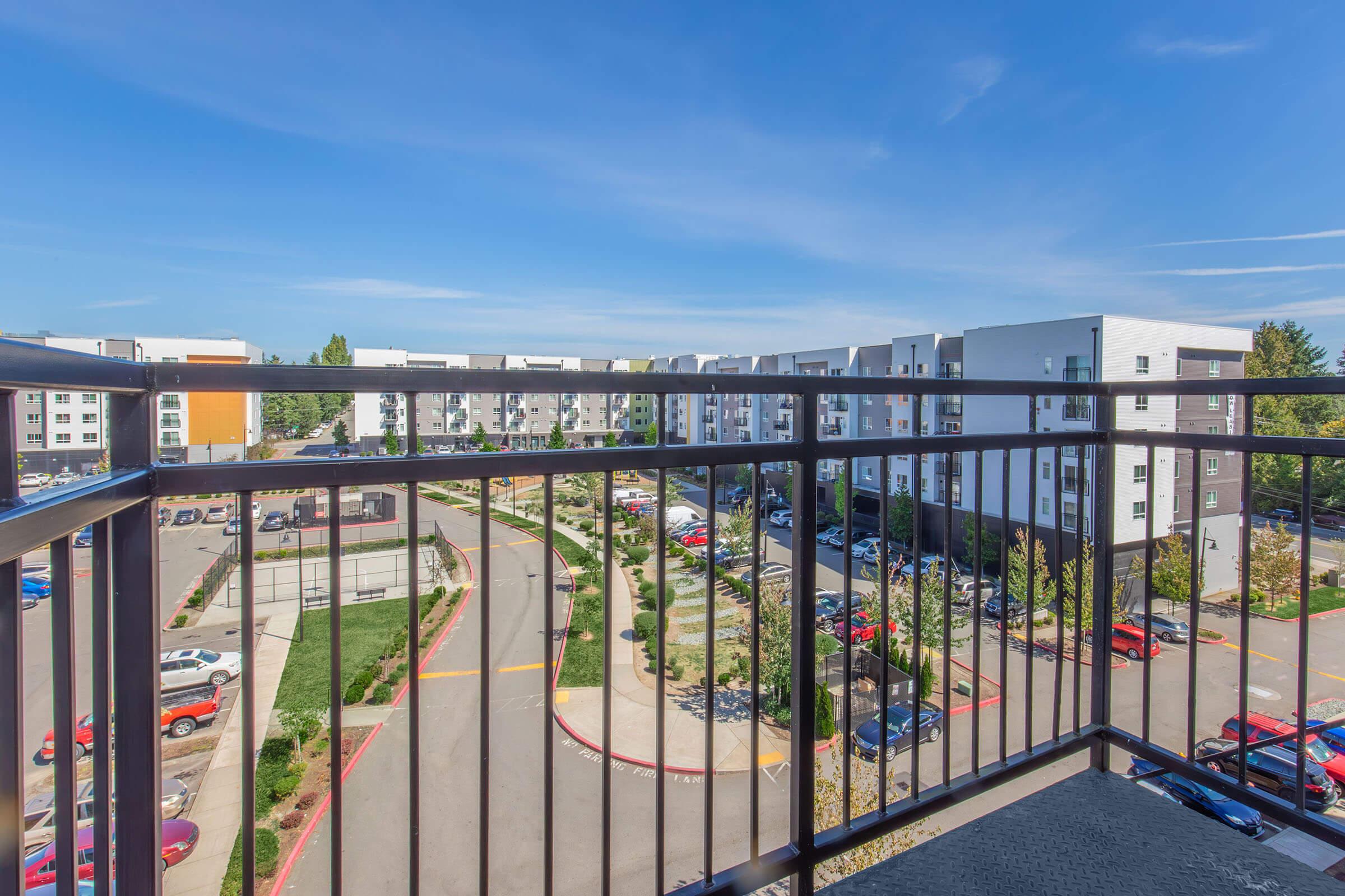 View from a balcony overlooking an apartment complex. The scene features several building structures, parked cars in a lot, and landscaped areas with greenery. The sky is clear with a few clouds, and the setting suggests a suburban environment.