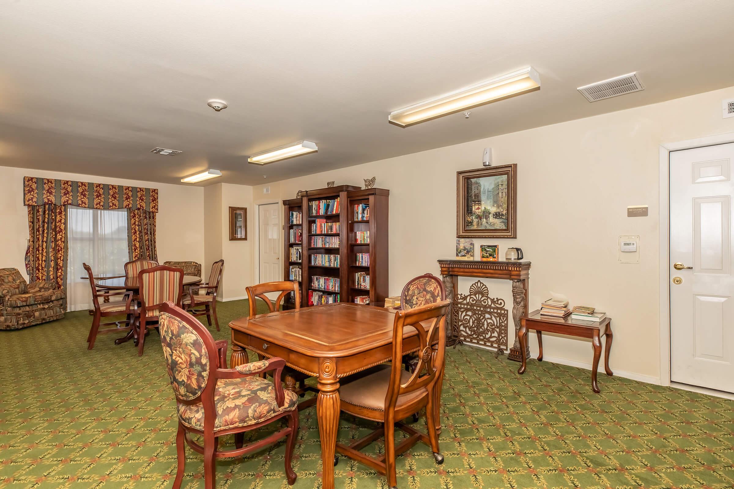 A cozy lounge area featuring a wooden bookshelf filled with books, a dining table set with chairs, and patterned soft furnishings. The walls are painted a soft color, and there are large windows with drapes, creating a warm and inviting atmosphere for relaxation and social gatherings.