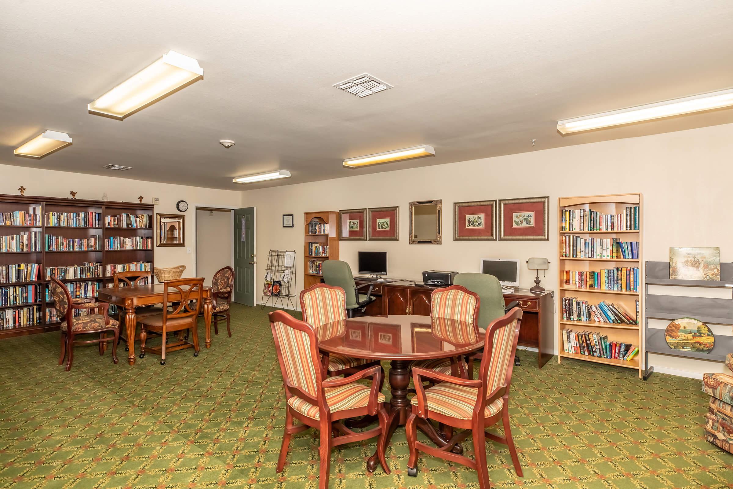 A cozy library or study area featuring bookshelves filled with various books. There are several round tables with chairs arranged for reading or studying. Two computers are placed on desks, and the room has a green carpet and warm lighting, creating an inviting atmosphere for relaxation and learning.