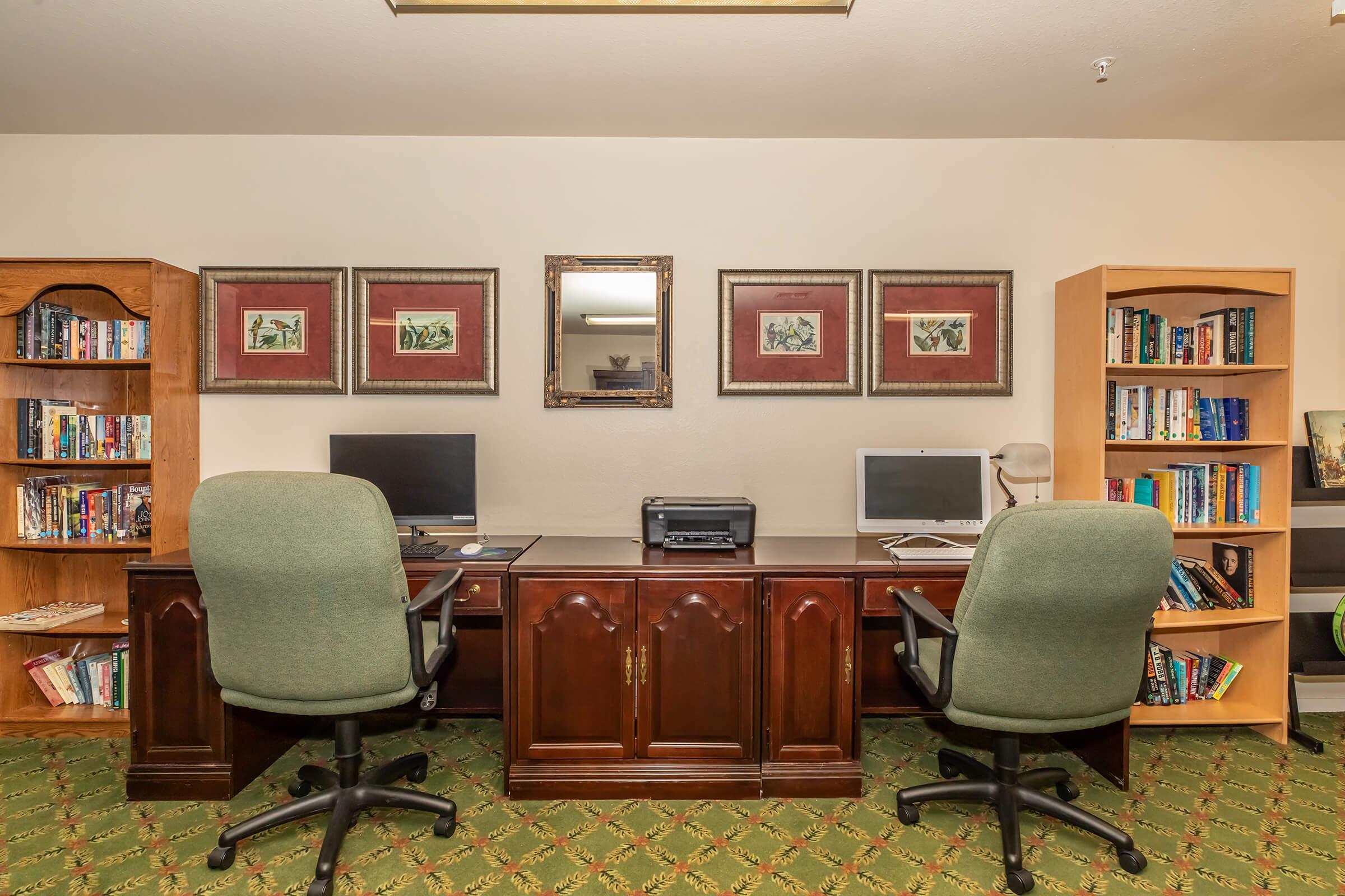 A cozy office space featuring two green office chairs positioned in front of wooden desks with computers. On the desks, there is a printer, and the walls are adorned with framed artwork. Bookshelves filled with various books are on either side, creating a warm and inviting atmosphere for work or reading.