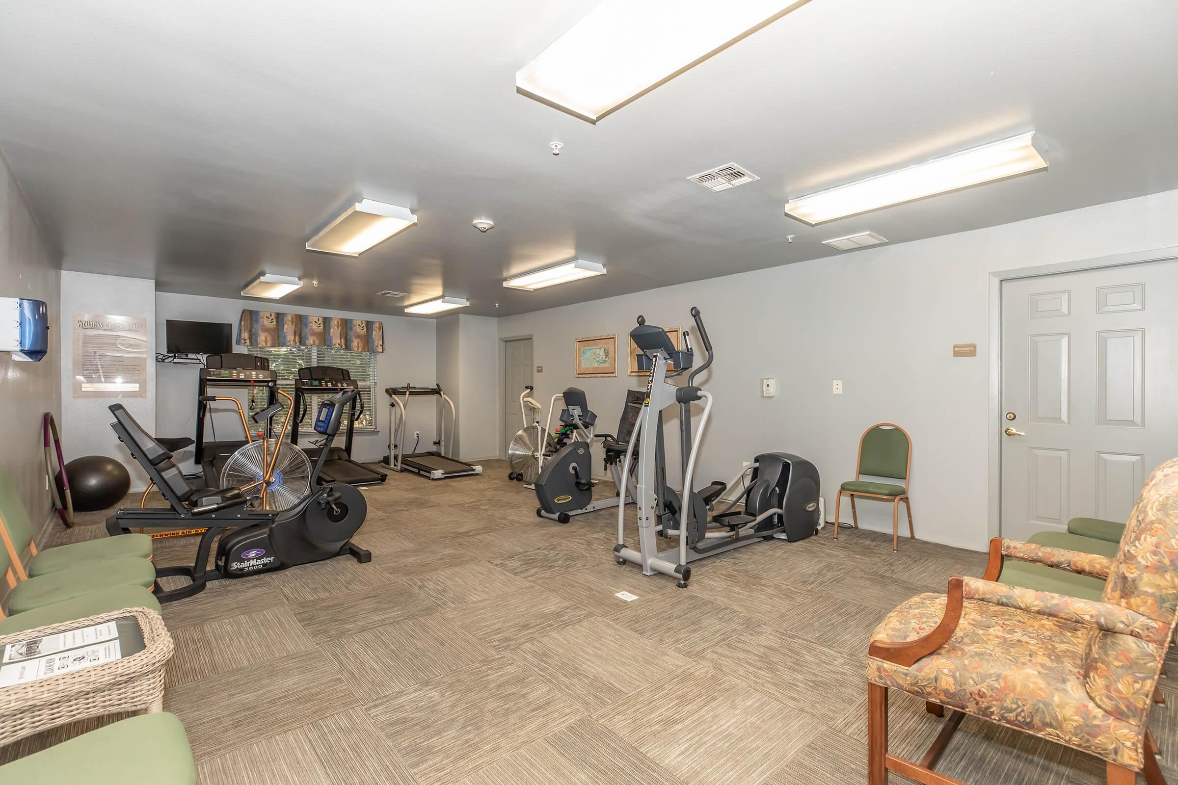 A fitness room featuring various exercise equipment including treadmills, stationary bikes, and an elliptical machine. The space has a few chairs and is well-lit with overhead lighting. A wall-mounted TV is visible, along with framed artwork and a welcoming atmosphere for exercise.