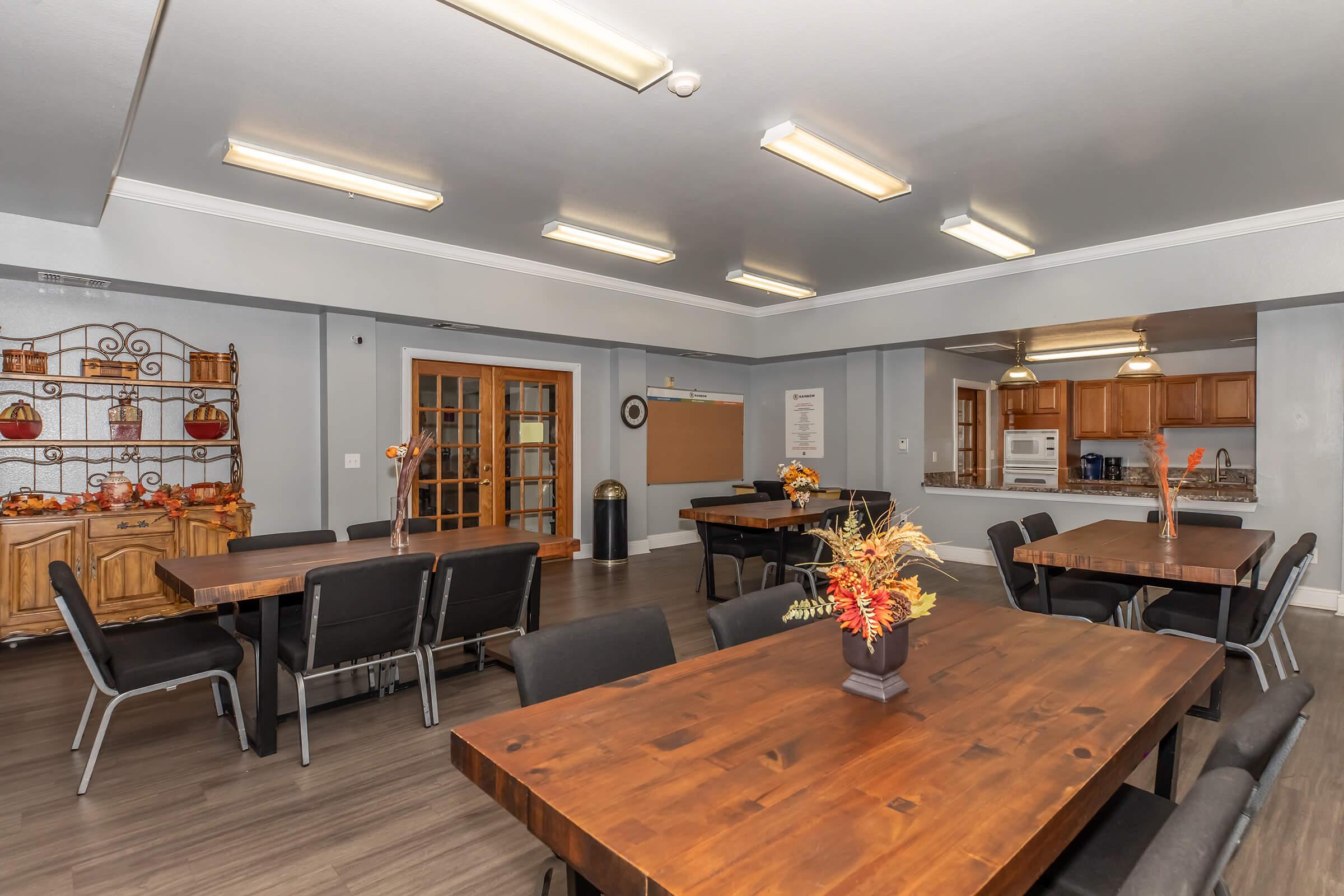 A spacious dining area featuring several large wooden tables with black chairs, decorated with flower vases. The room has soft lighting and a kitchenette partially visible in the background, along with a wall shelf displaying decorative items and seasonal decor.