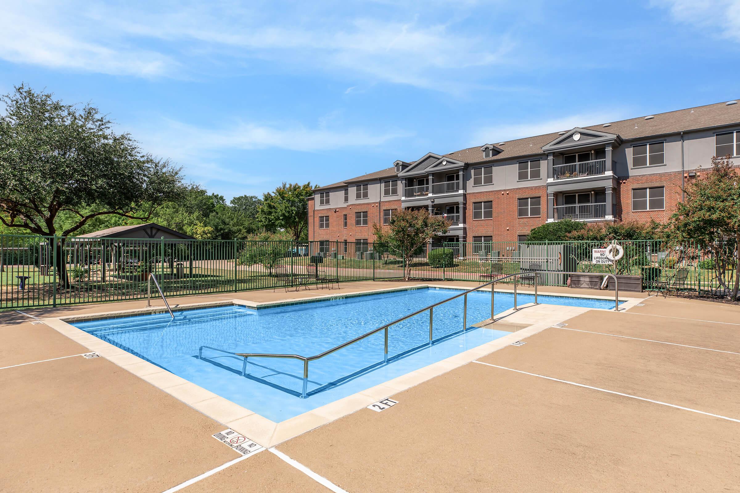 A clear swimming pool surrounded by a tan deck and green lawn. In the background, a three-story apartment building with balconies can be seen, along with trees and a small pavilion. The sky is bright and blue, creating a sunny atmosphere.