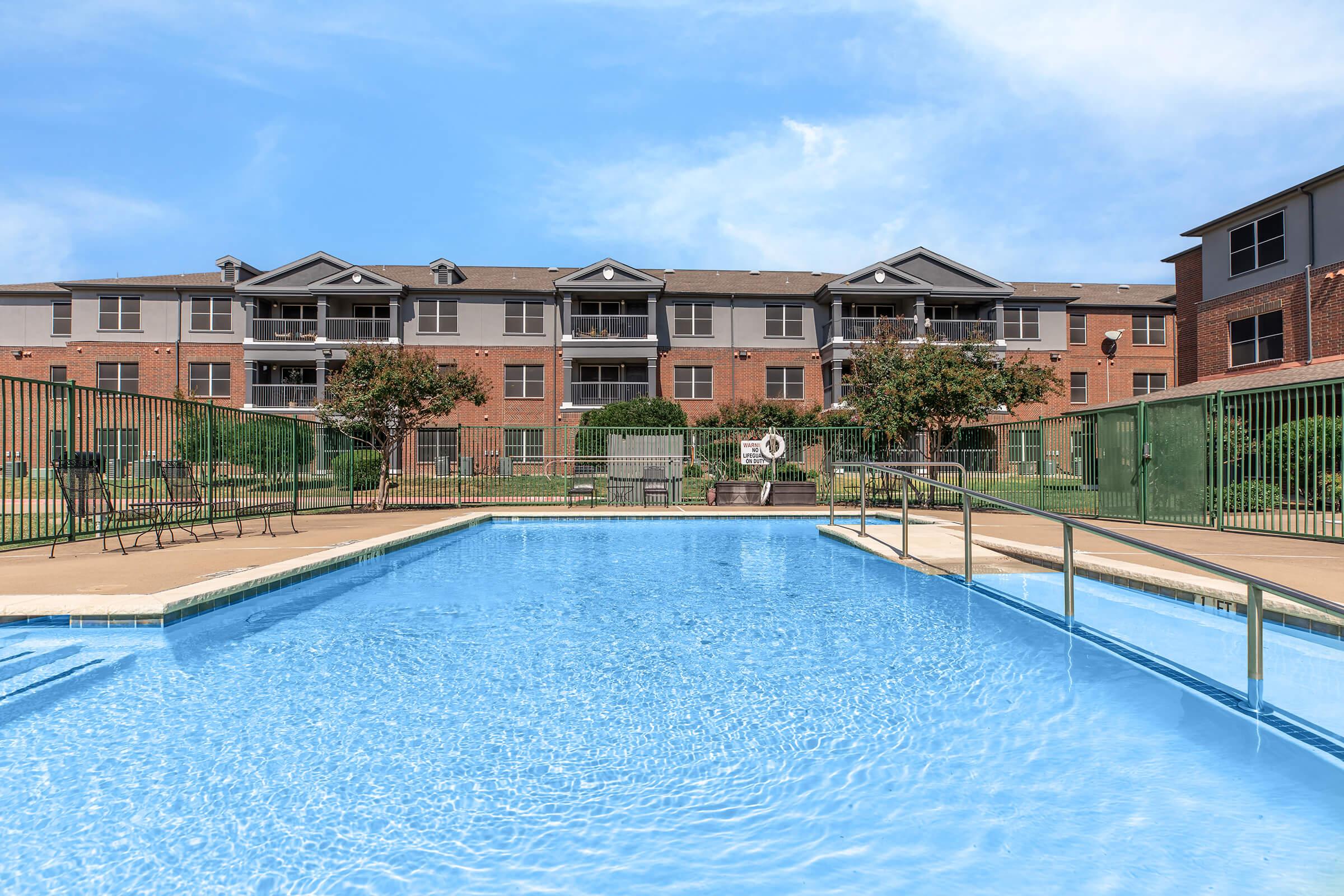 A clear blue swimming pool in the foreground with a fenced area. In the background, there are red brick apartment buildings and green foliage. The sky is bright with a few clouds visible. Poolside chairs and tables are arranged near the water.