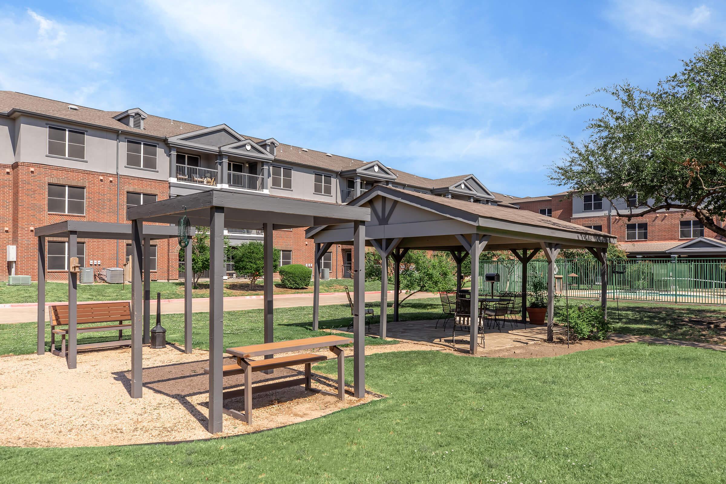 A grassy area featuring two wooden gazebos with benches, surrounded by a landscaped environment. In the background, there's a multi-story brick apartment building. Clear blue sky overhead creates a bright and inviting atmosphere.