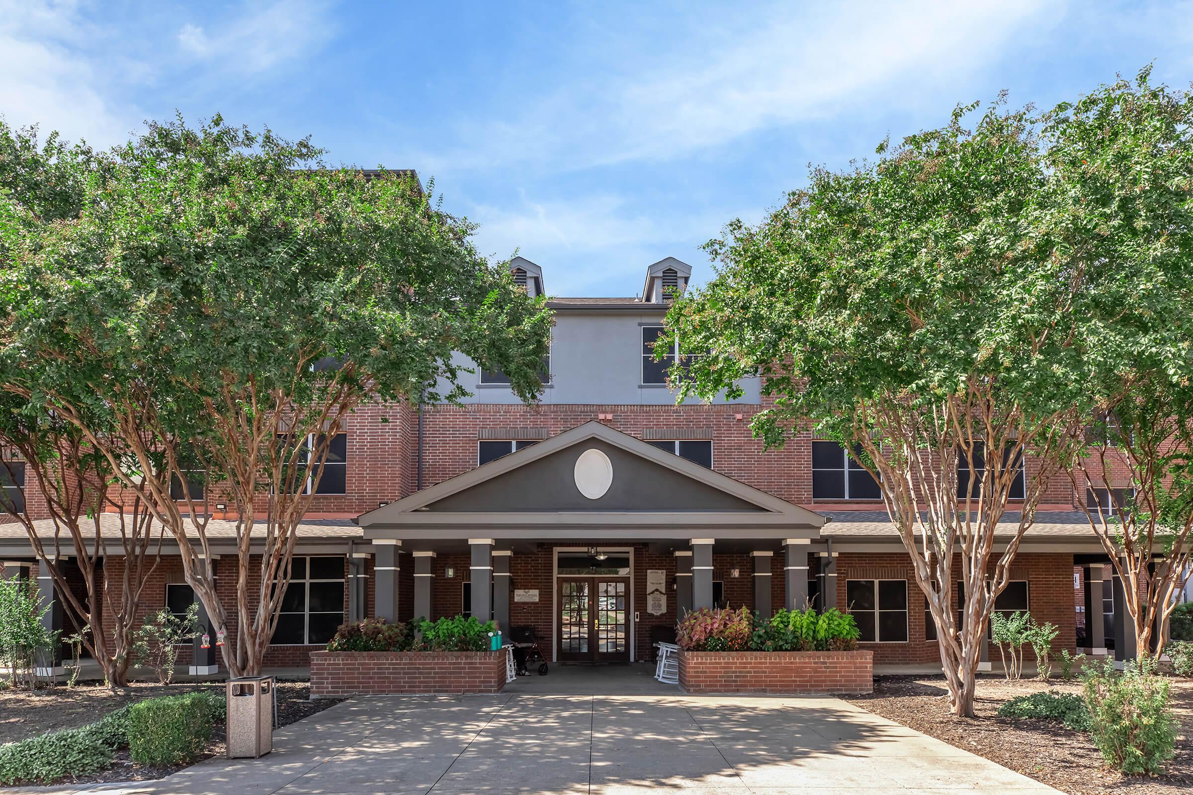 A brick building with a large entrance, featuring a covered porch and decorative features. Flanking the entrance are several trees and landscaped areas. The sky above is clear with a few clouds. The scene is well-lit, suggesting a welcoming environment.