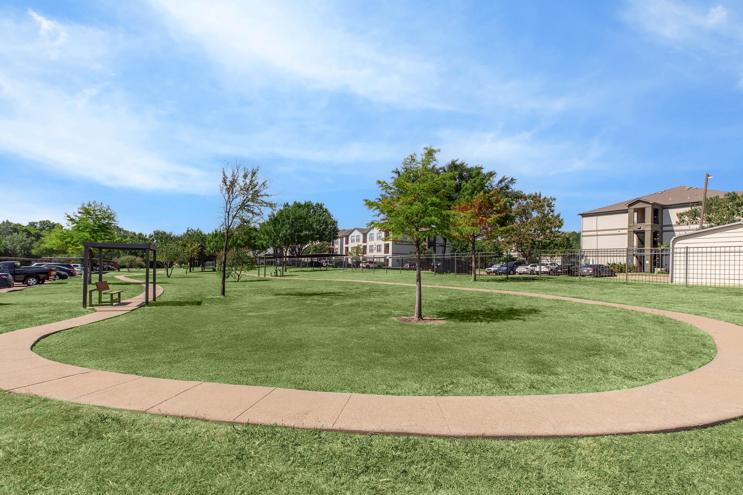 A spacious park area featuring a neatly manicured green lawn, a winding pathway, and a few young trees. In the background, there are residential buildings and parked cars, with a clear blue sky overhead. The setting offers a peaceful atmosphere for leisure activities.