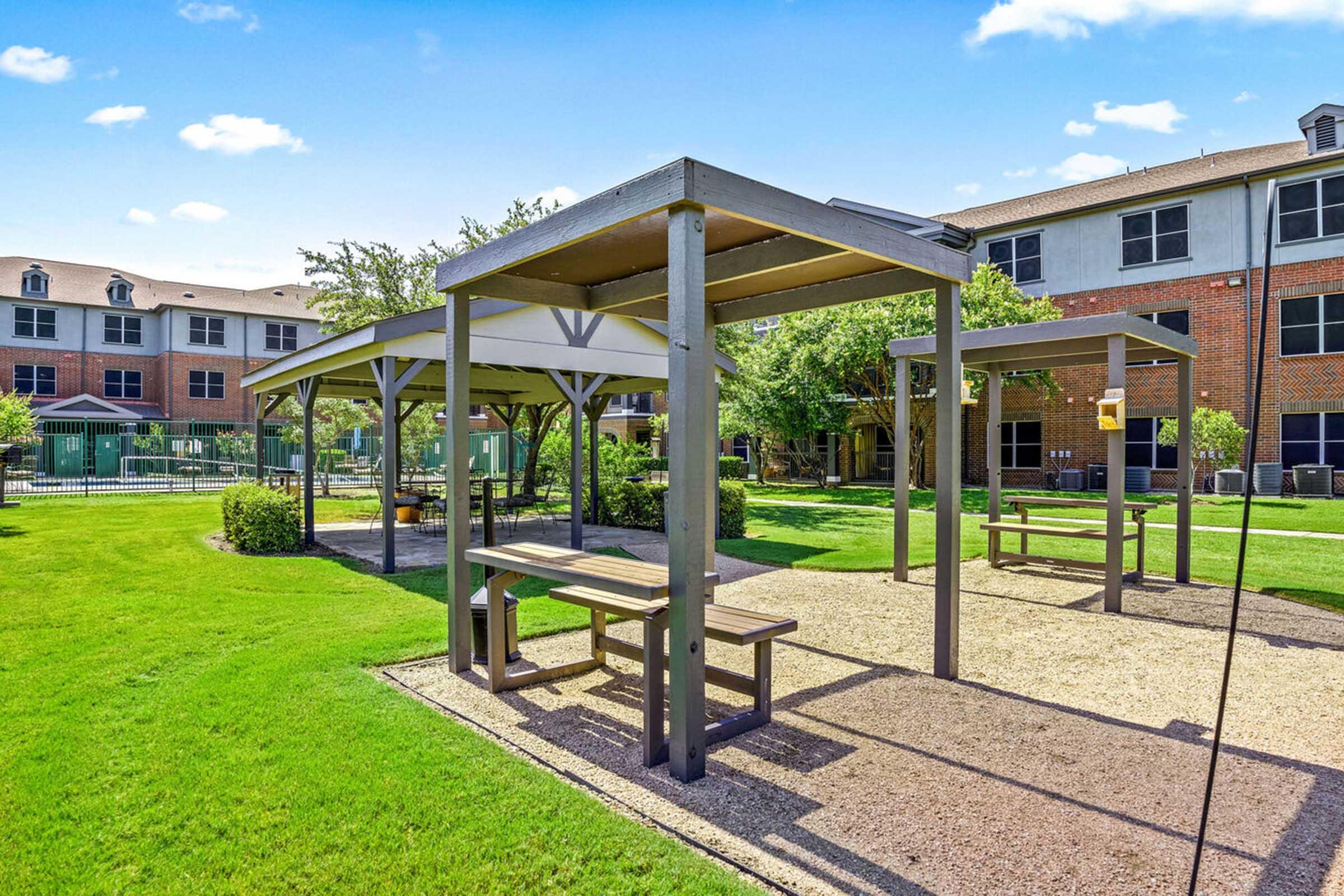 A sunny outdoor space featuring several shaded picnic areas with wooden benches under metal canopies.