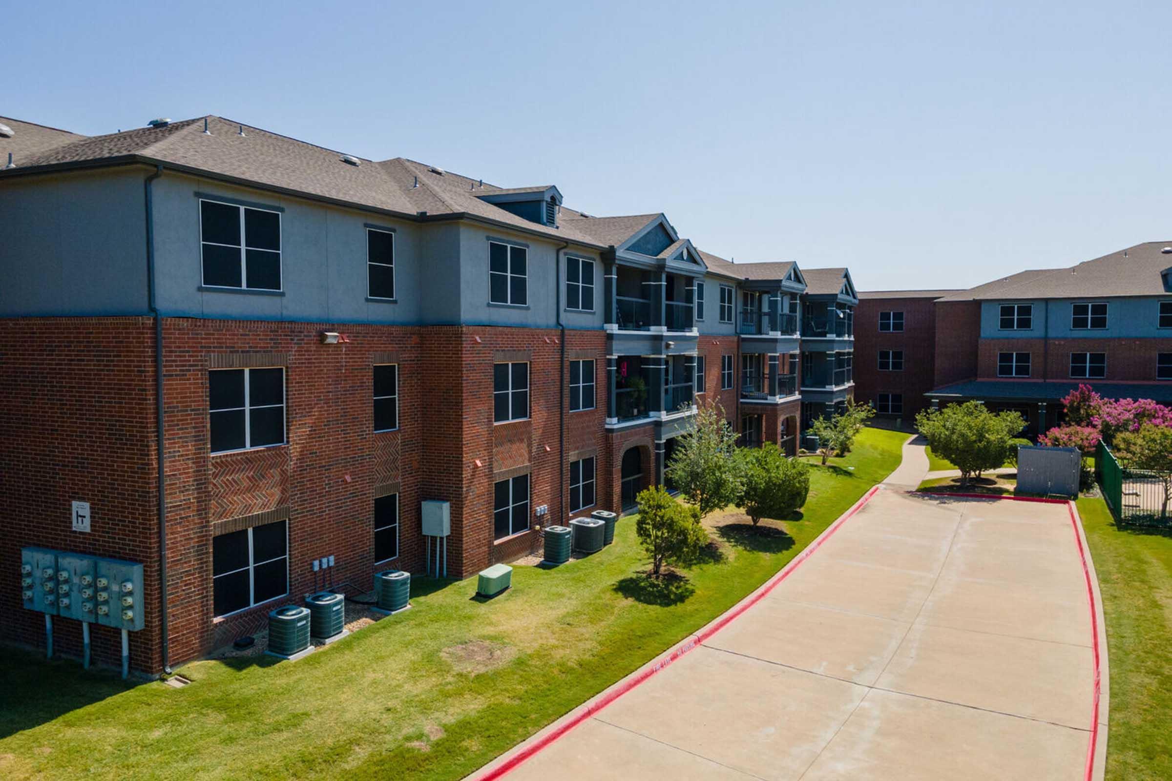 A modern apartment complex featuring brick and blue exterior walls, surrounded by well-maintained lawns and landscaping. T