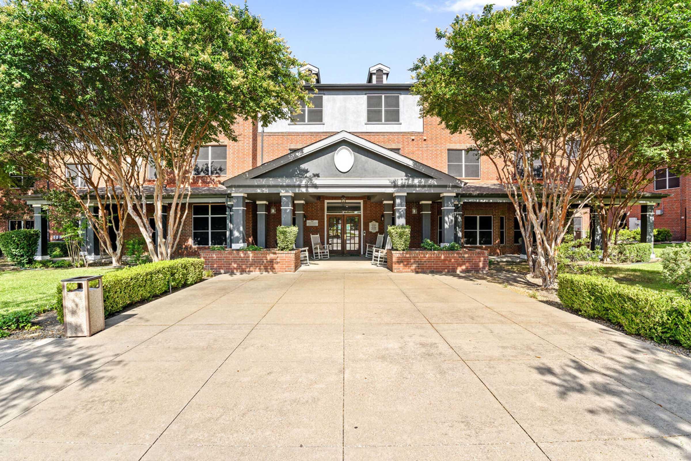 A well-maintained brick building with a large entrance, flanked by two trees. The pathway leads to the entrance, which features a covered porch and rocking chairs.